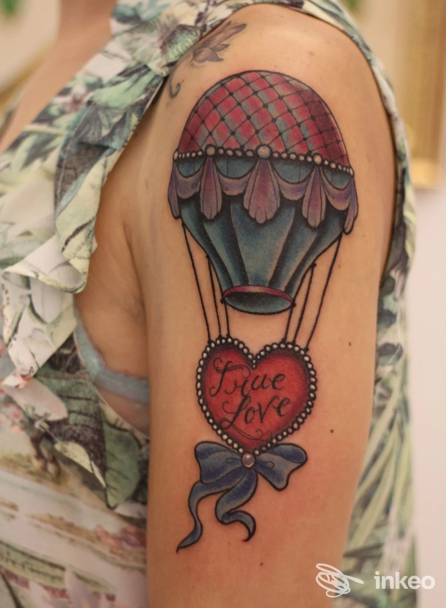 Braccio visibile con tatuaggio realistico: mongolfiera colorata dai toni vividi, con cuore rosso contenente scritta love e fiocco blu, su cute sana.