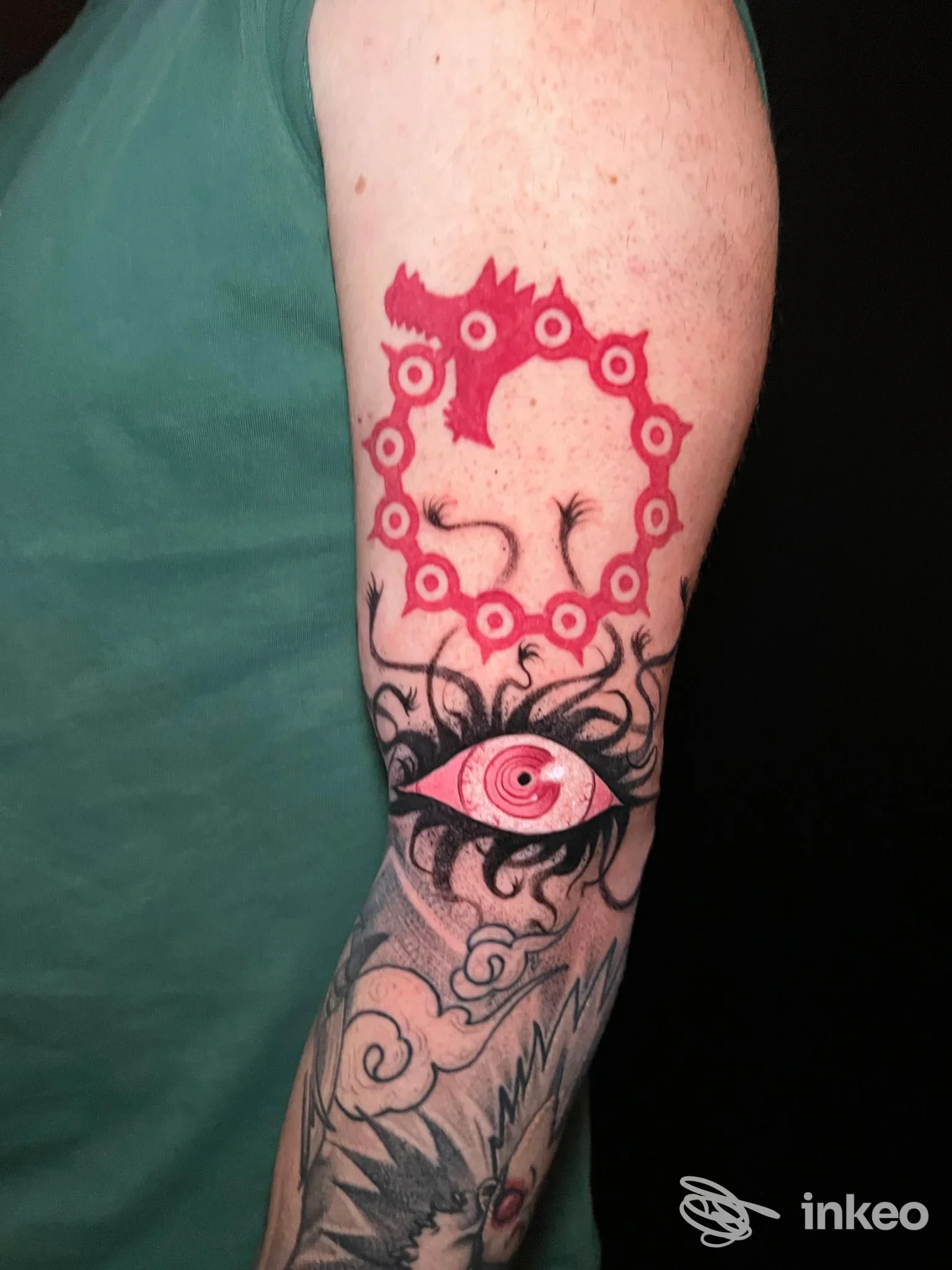 Tattoo sul braccio raffigurante un occhio circondato da una catena rossa, con elementi geometrici e nuvole grigie sullo sfondo.