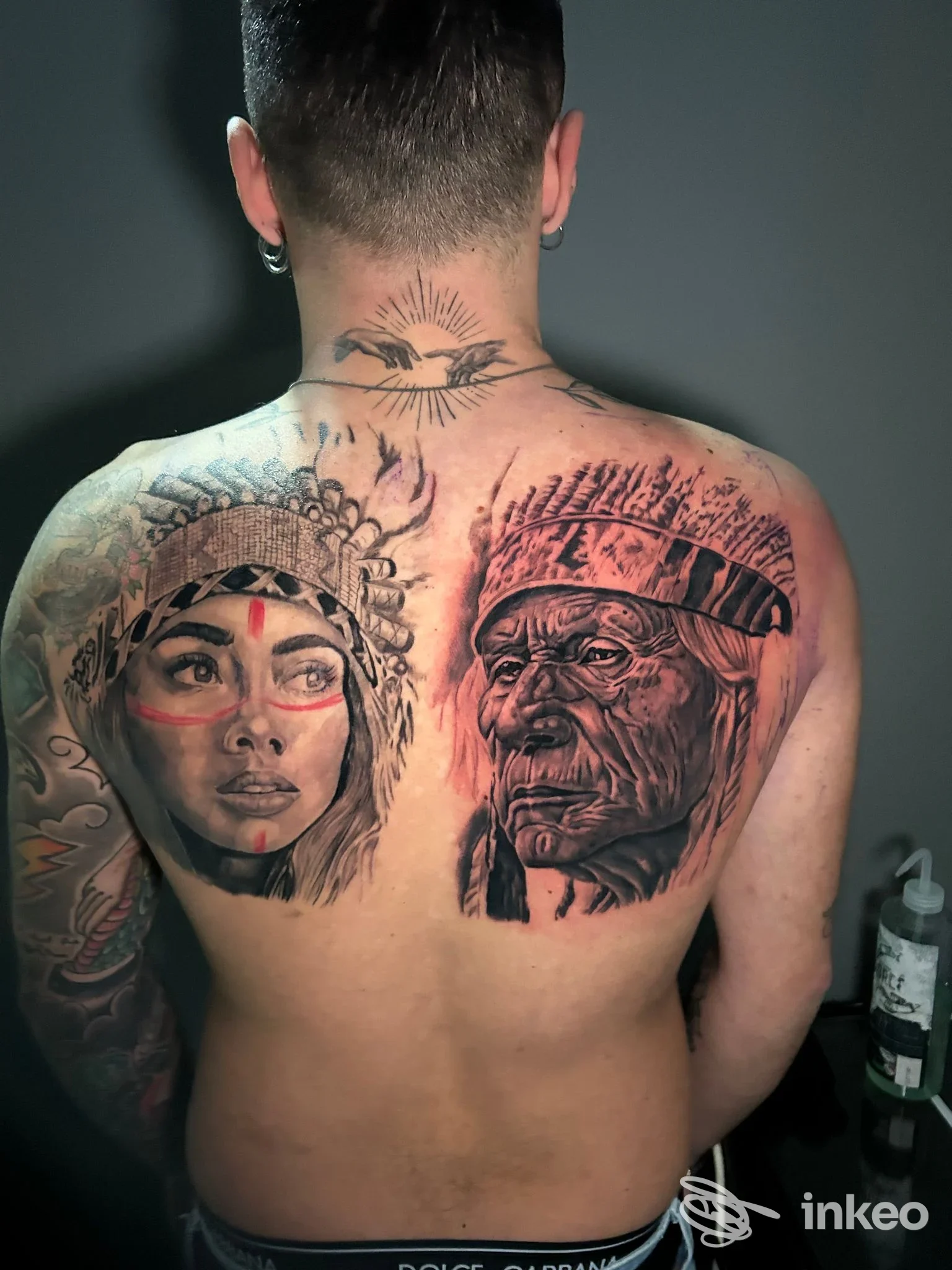Schiena di un uomo con due tatuaggi: un volto nativo americano a sinistra e un vecchio uomo a destra, in bianco e nero, tatuaggi ben definiti sul dorso. luce fredda.