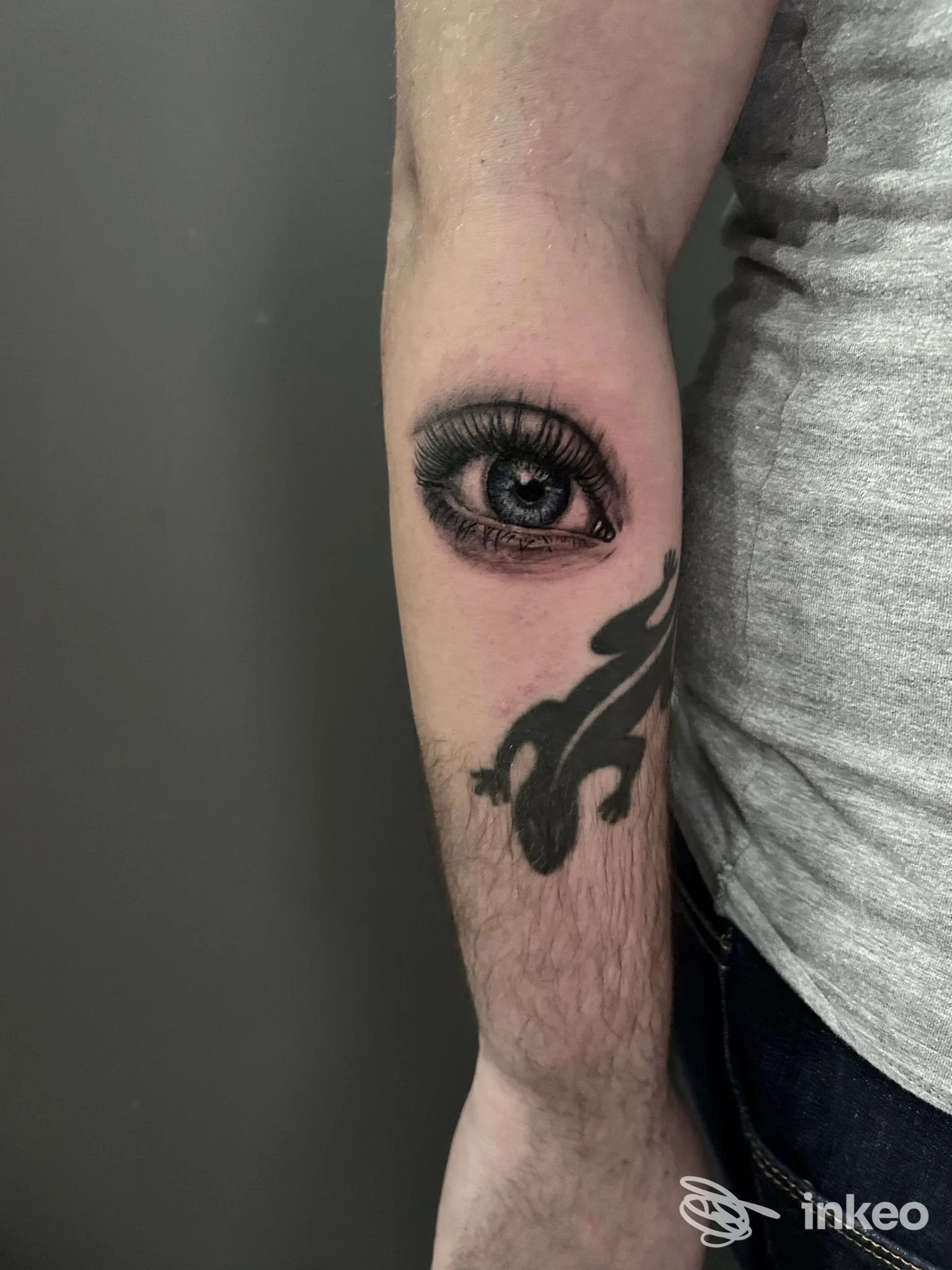 Tattoo realistico di un occhio singolo sul braccio, accompagnato da un motivo floreale scuro.