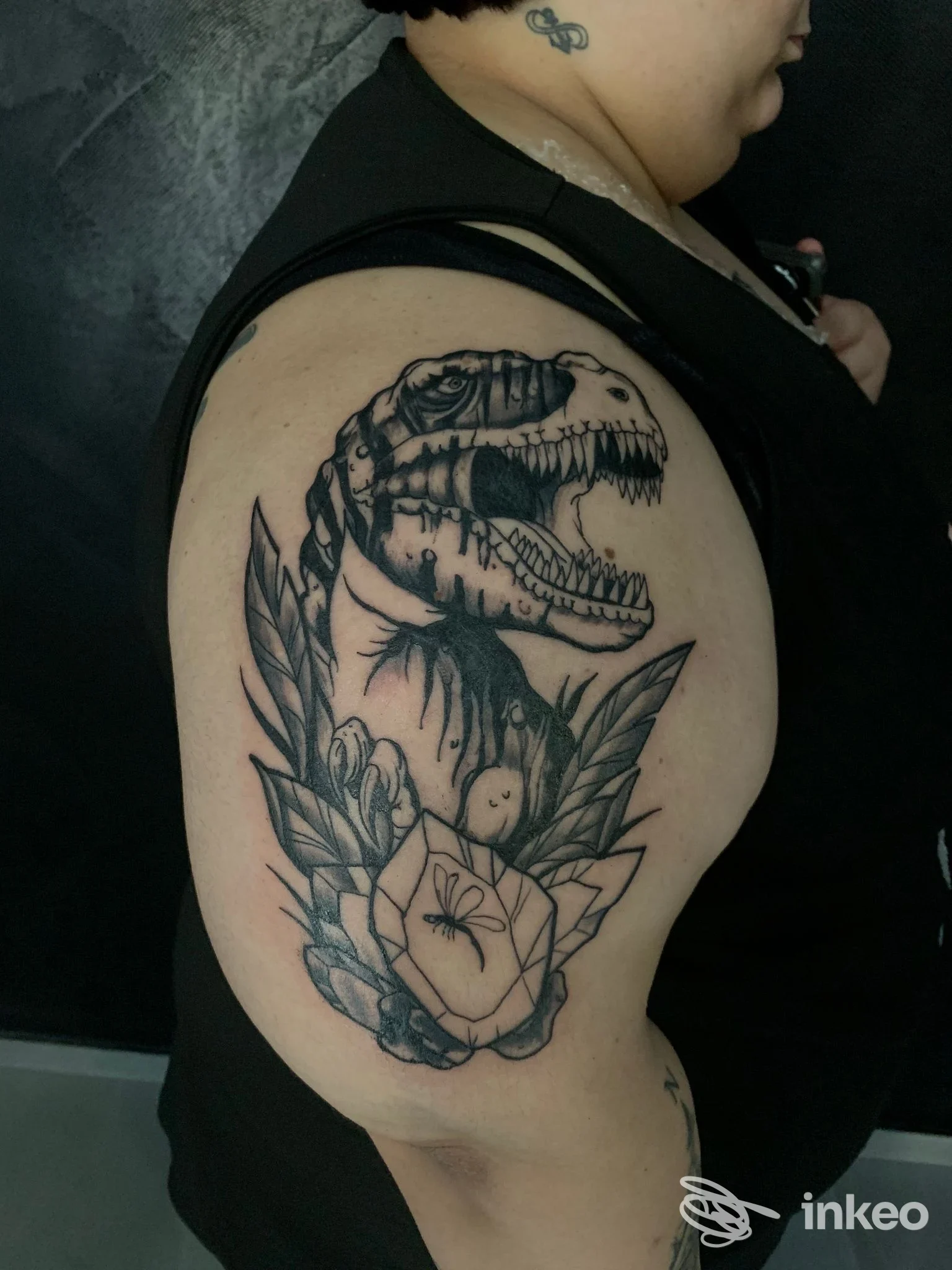 Tatuaggio realistico di dinosauro T-Rex sulla spalla di una persona, con fiori e foglie decorative attorno; realizzo su pelle, nessun overlay social o schizzo preparatorio visibile.