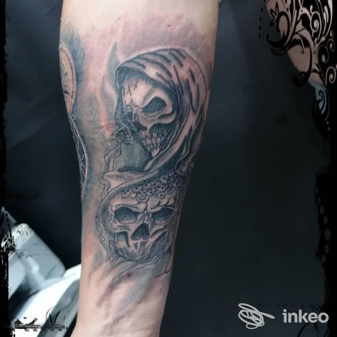 Tatuaggio in bianco e nero sul braccio raffigurante una figura incappucciata e teschi, realizzato con tonalità grigie su pelle, visibile in un contesto di studio.