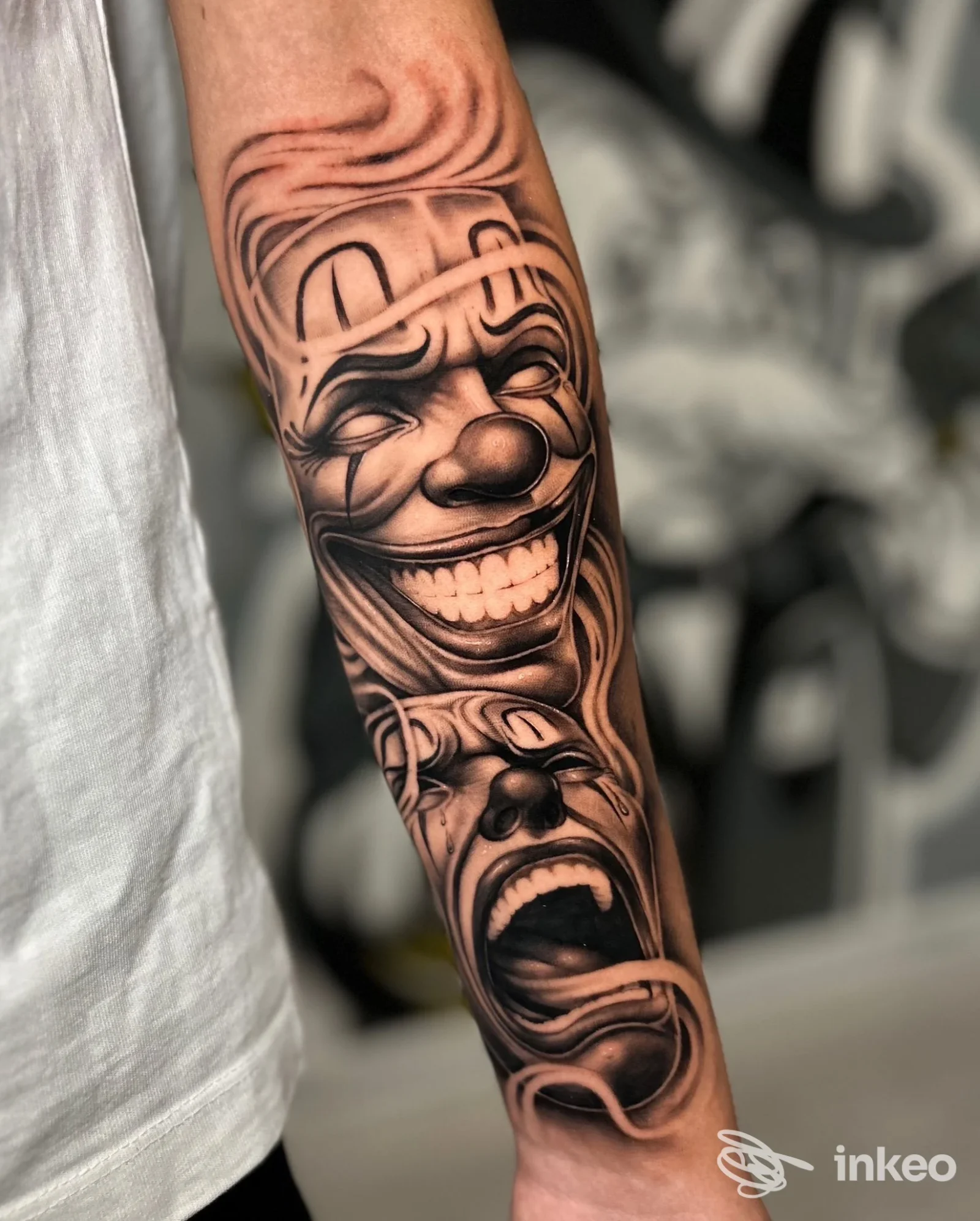 Tattoo visibile sullavambraccio raffigurante due maschere teatrali: una sorridente e una urlante, eseguito su pelle reale; colori in bianco e nero con tratto definito.