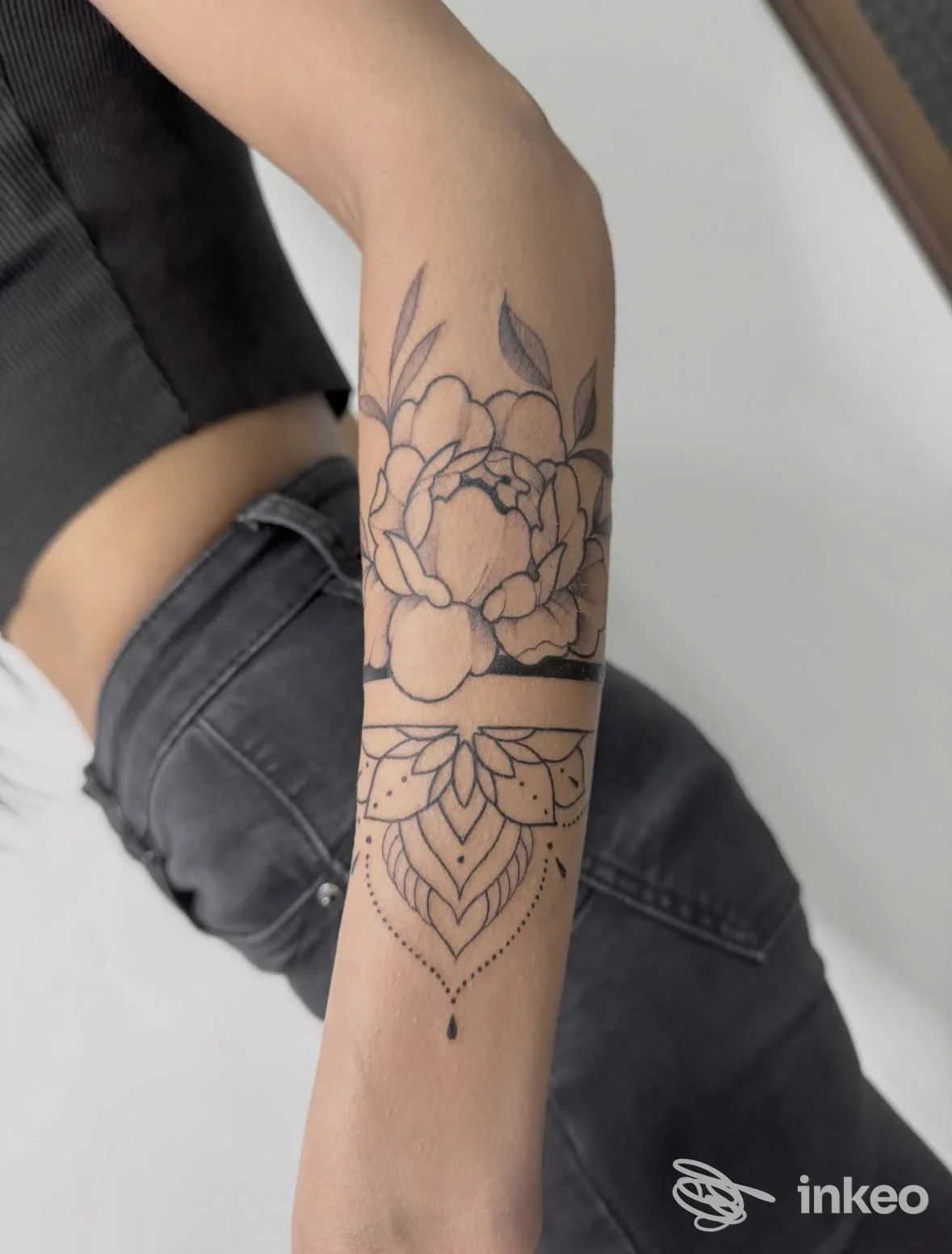 Avambraccio visibile con tatuaggio floreale in linee nere su pelle, motivo decorativo con pendente sotto la sezione centrale. foto laterale senza volto visibile.