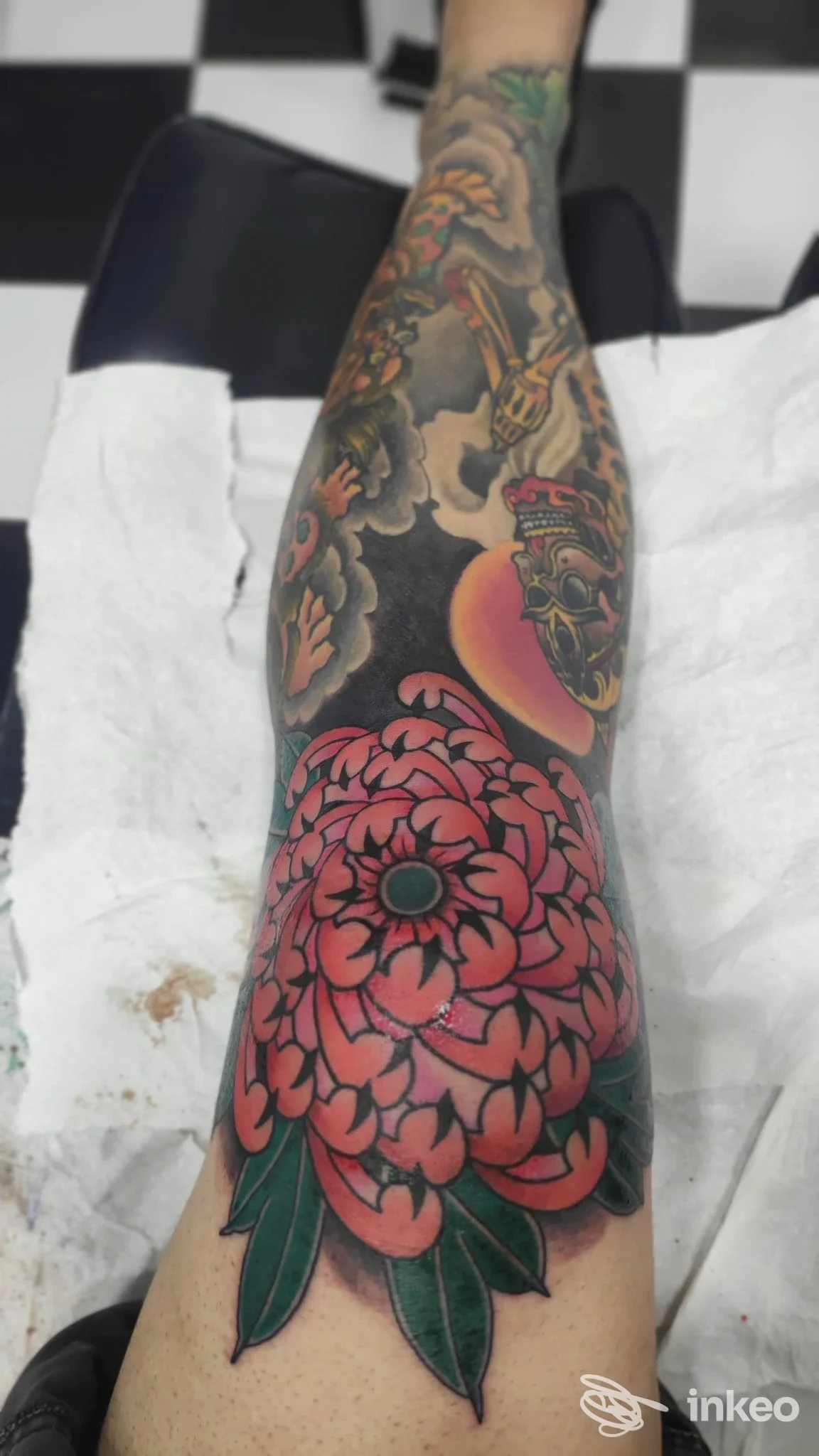Avambraccio/caviglia visibile con tatuaggio grande a tema fiore rosa paesaggistico, petali evidenti e foglie verdi su sfondo scuro.