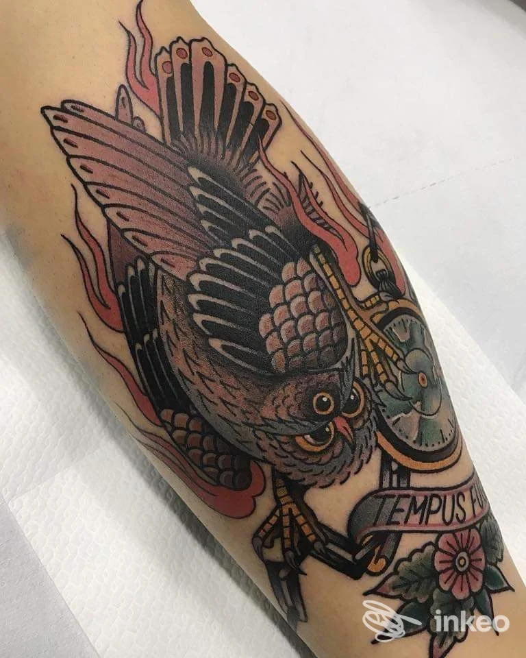 Tattoo su braccio raffigurante un falco con ali dettagliate, pennacchi e fiamme, accompagnato da nastrino e fiore, colore vivace su pelle. Napoli.