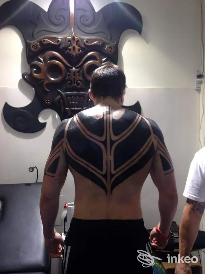 Un uomo visto di schiena mostra un grande tatuaggio geometrico nero sulla schiena, stile tribale, realizzato sulla pelle; l’ambientazione è uno studio con decorazioni murali; colore dominante nero.