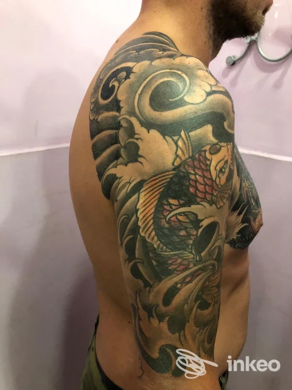 Tattoo sul braccio e spalla sinistra di un uomo, stile tradizionale/girevole con nuvole e pesce, colori scuri e linee nette.