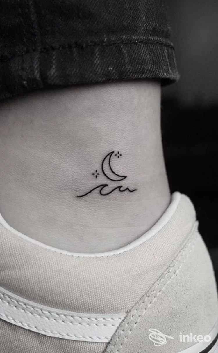 Tatuaggio minimale su caviglia: luna crescente e linee che sembrano onde, eseguito sulla pelle chiara, con jeans scuri che sfiorano la zona. studio Mike Biscuit, Parete.