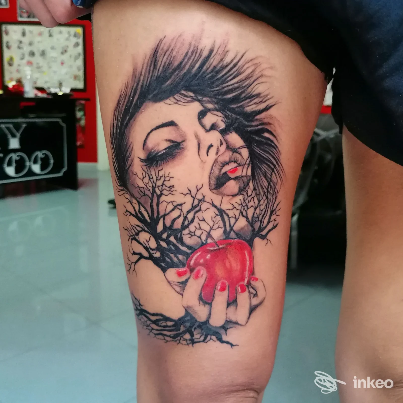 Tattoo raffigurante un volto femminile con rami e una mela rossa tra le mani, tatuaggio su stinco. colori essenziali in bianco e nero con accento rosso sulla mela.