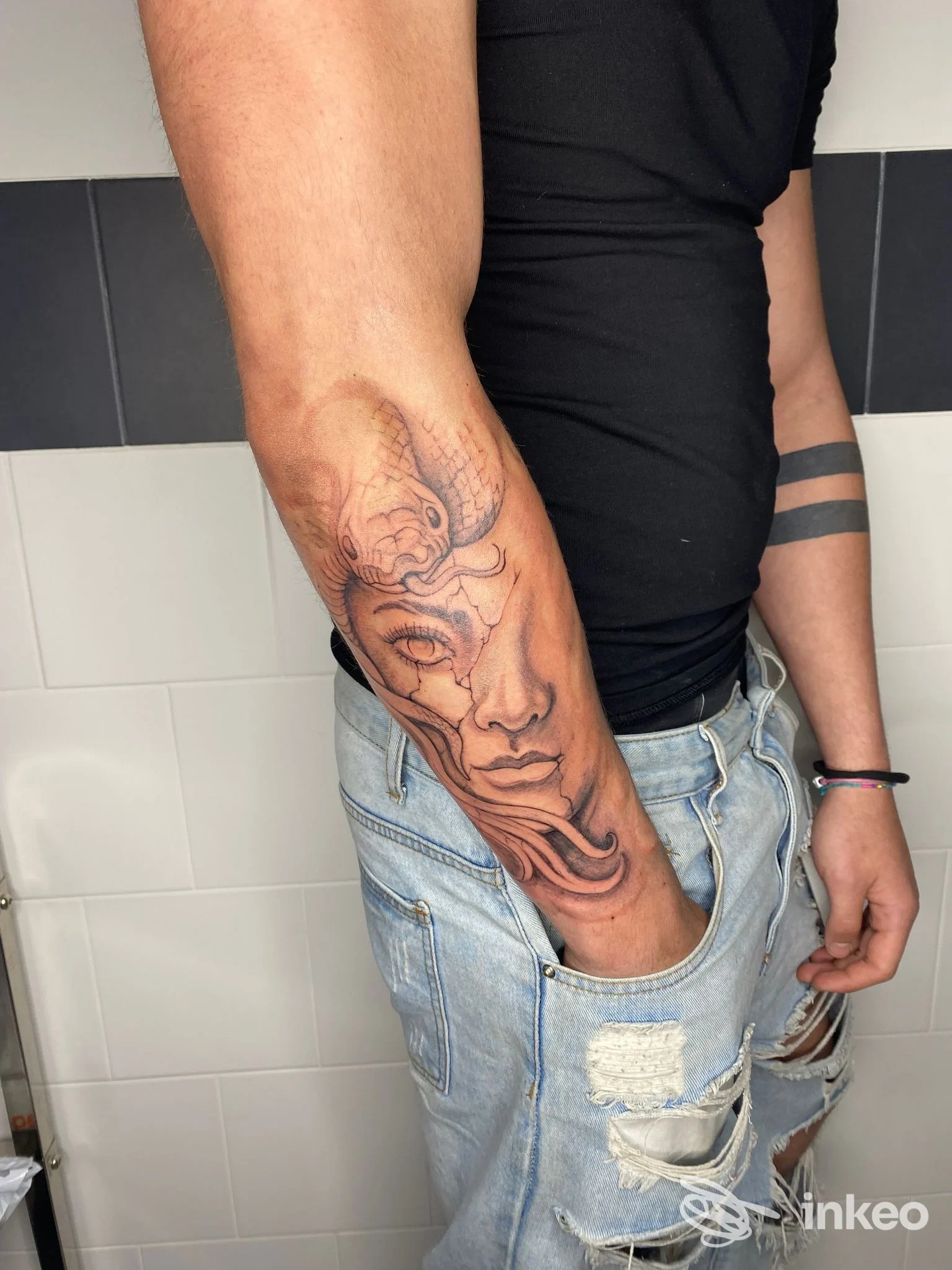 Tattoo sull’avambraccio di una persona, realizzato presso INKiostro Tattoo Art a Mugnano del Cardinale, raffigurante un volto stilizzato con linee nette; l’immagine mostra braccio con jeans strappati.