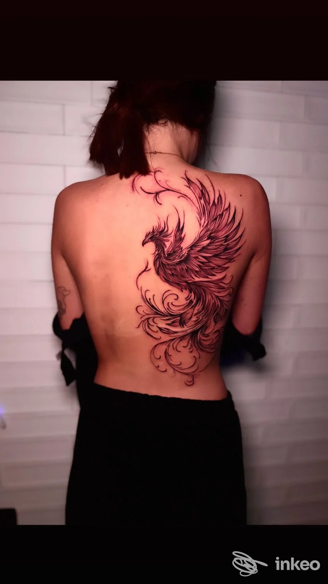 Tattoo reale sulla schiena di una persona: una fenice nera e rossa con linee eleganti, scatto verticale; studio Blackout Ink Tattoo Studio, Quarto.