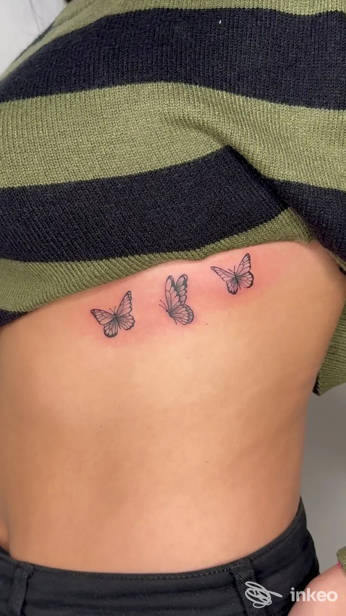 Tre farfalle tatuate lungo la costola di una persona, visibili sotto una t-shirt sollevata, senza elementi di social overlay.