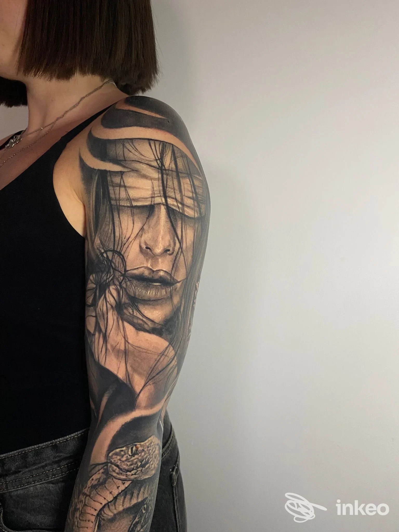 Tattoo realistico su braccio di una persona, ritratto parziale integrato con motivi decorativi, in tonalità nero/grigio; nessun volto intero visibile.