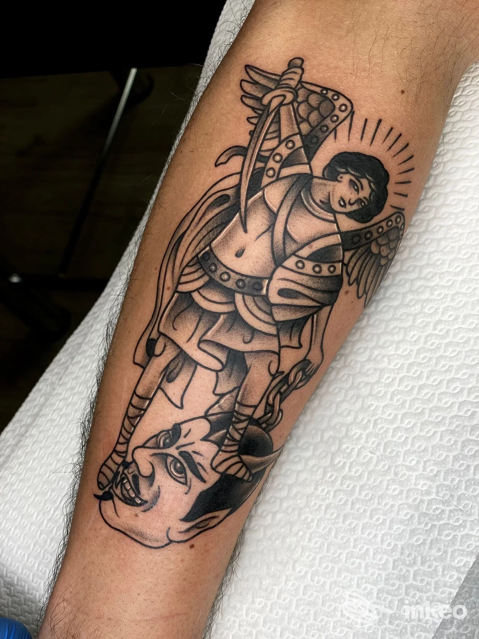 Tattoo sull'avambraccio raffigurante un angelo con spada che solleva una creatura demoniaca, disegno in stile linework tradizionale, pelle chiara.