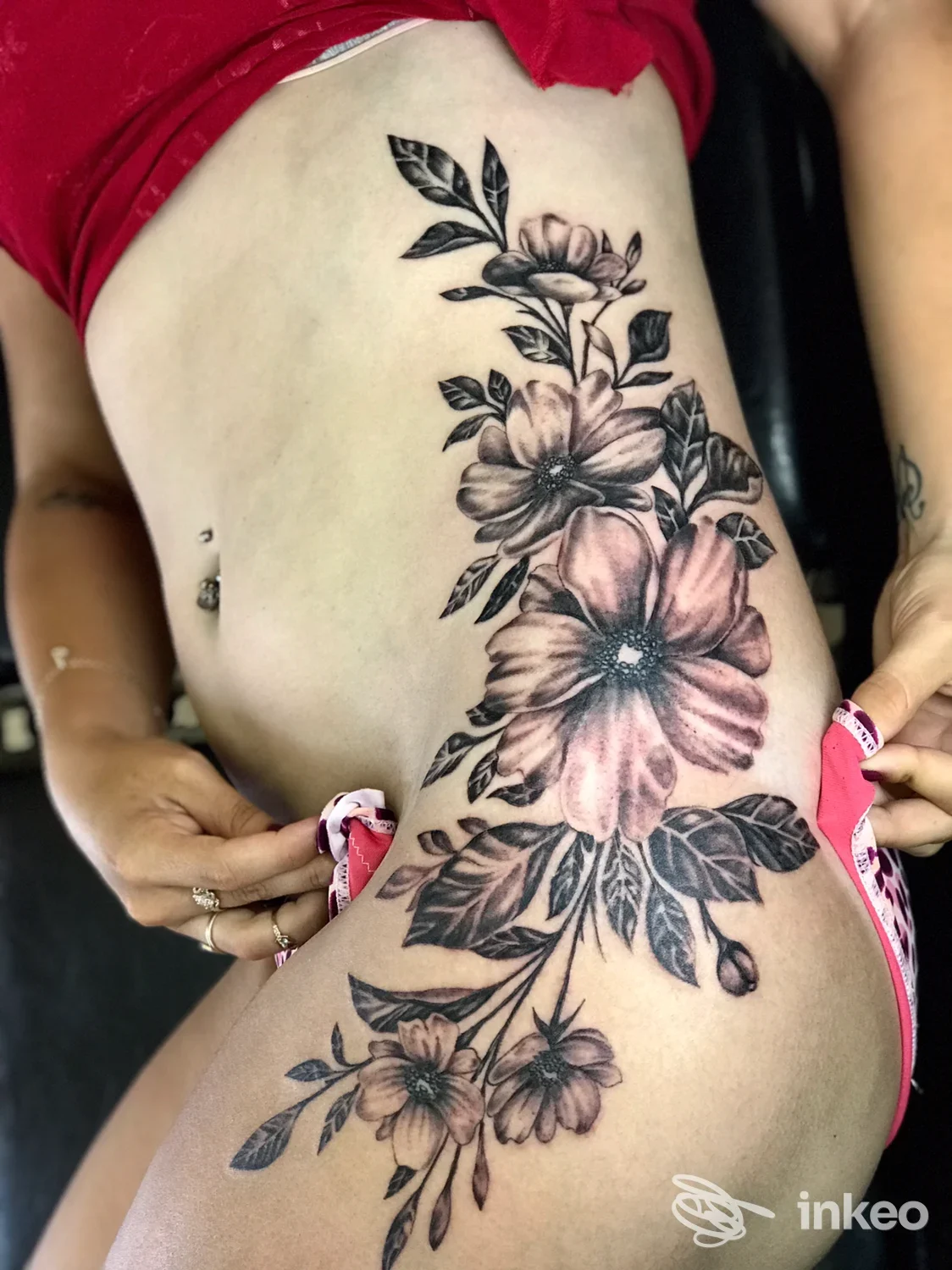 Tattoo floreale visibile sul fianco e sull’addome di una persona, colori rosa e grigio con foglie verdi, in posa laterale.