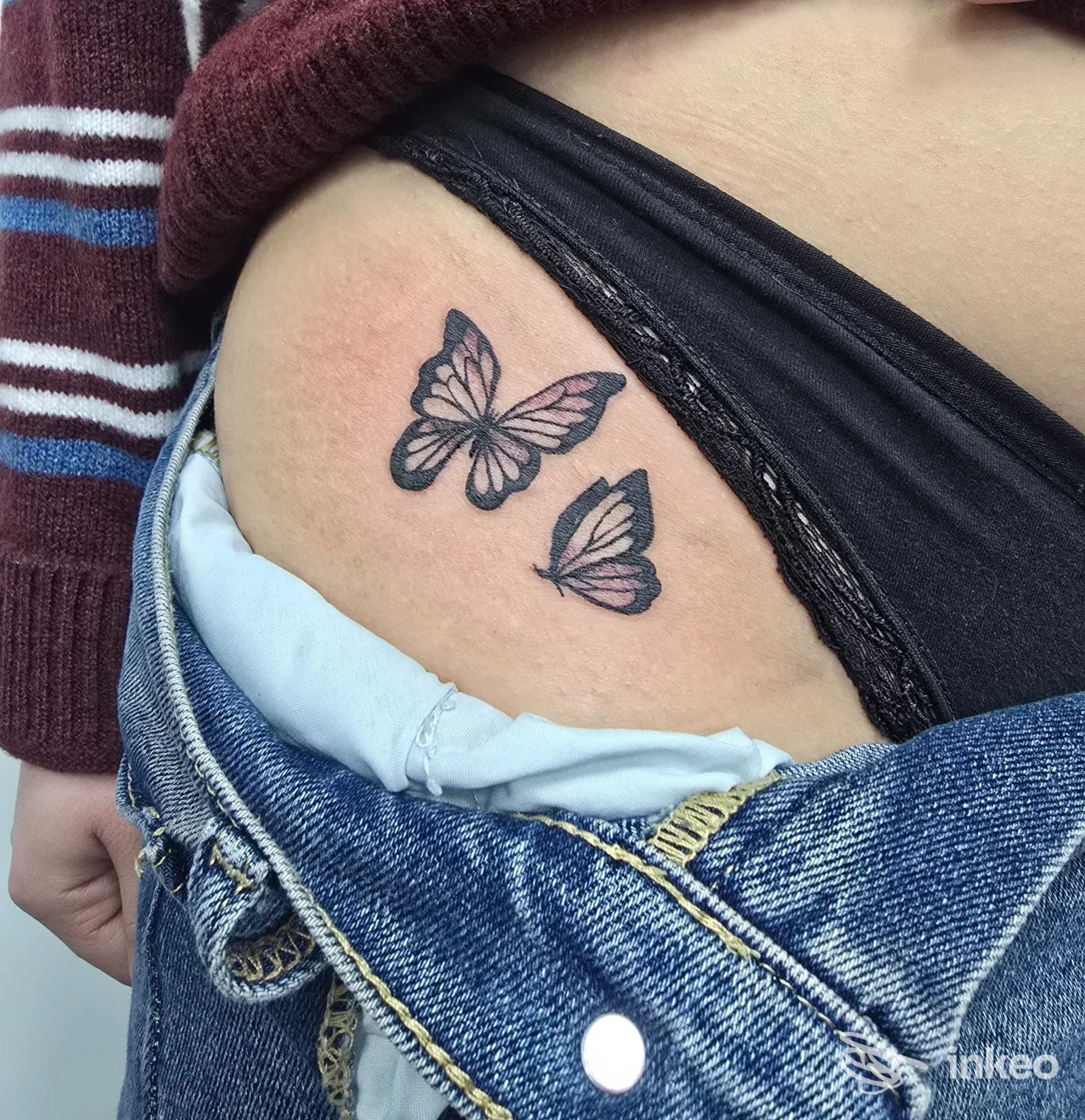 Piccolo tatuaggio di due farfalle poste sulla pelle, visibile sull’anca/fianco: colori neri con sfumature rosa, abbinato a jeans e maglia.