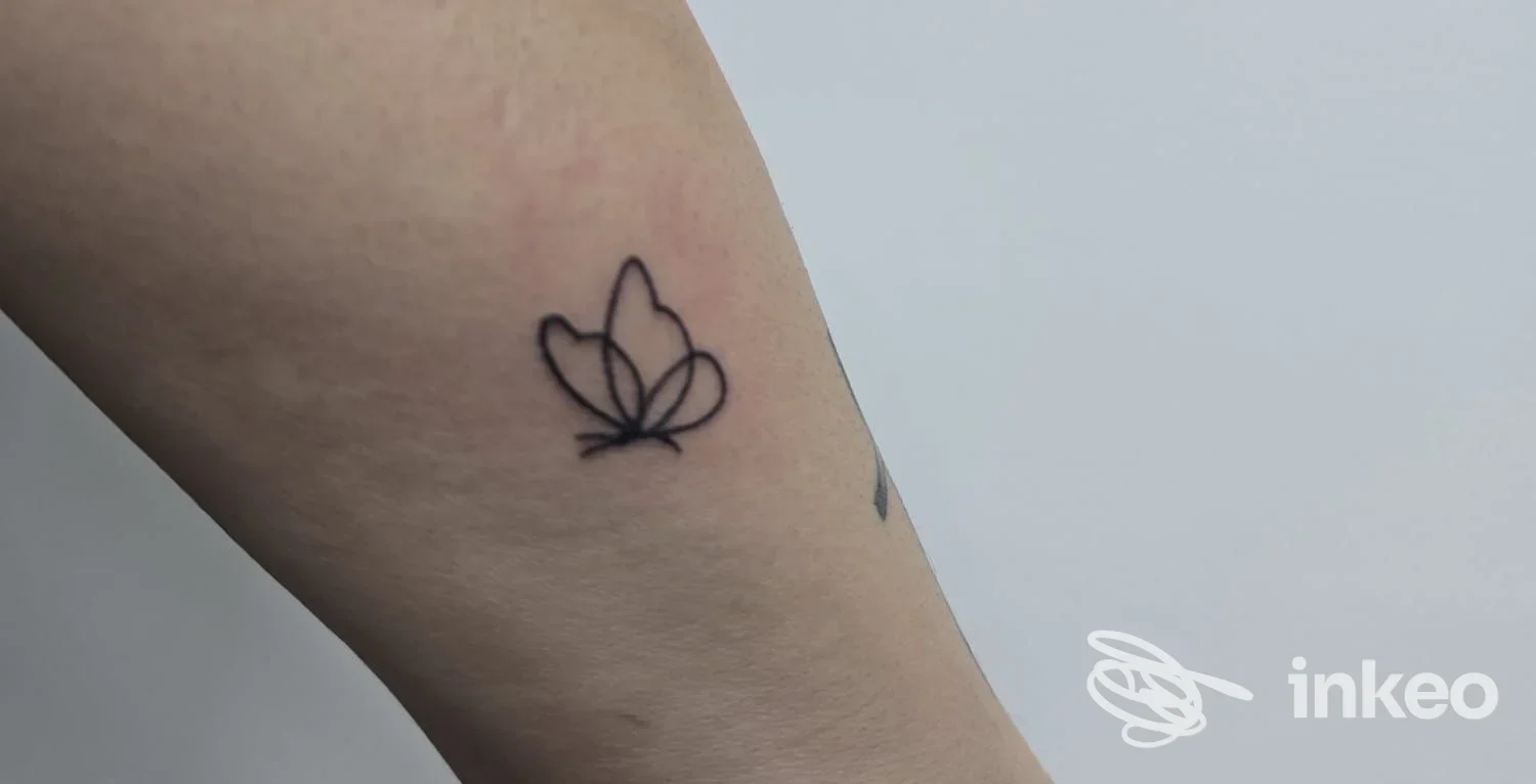 Tatuaggio reale visibile sull’avambraccio: piccolo disegno stilizzato a forma di farfalla, tatto lineare in nero su pelle.