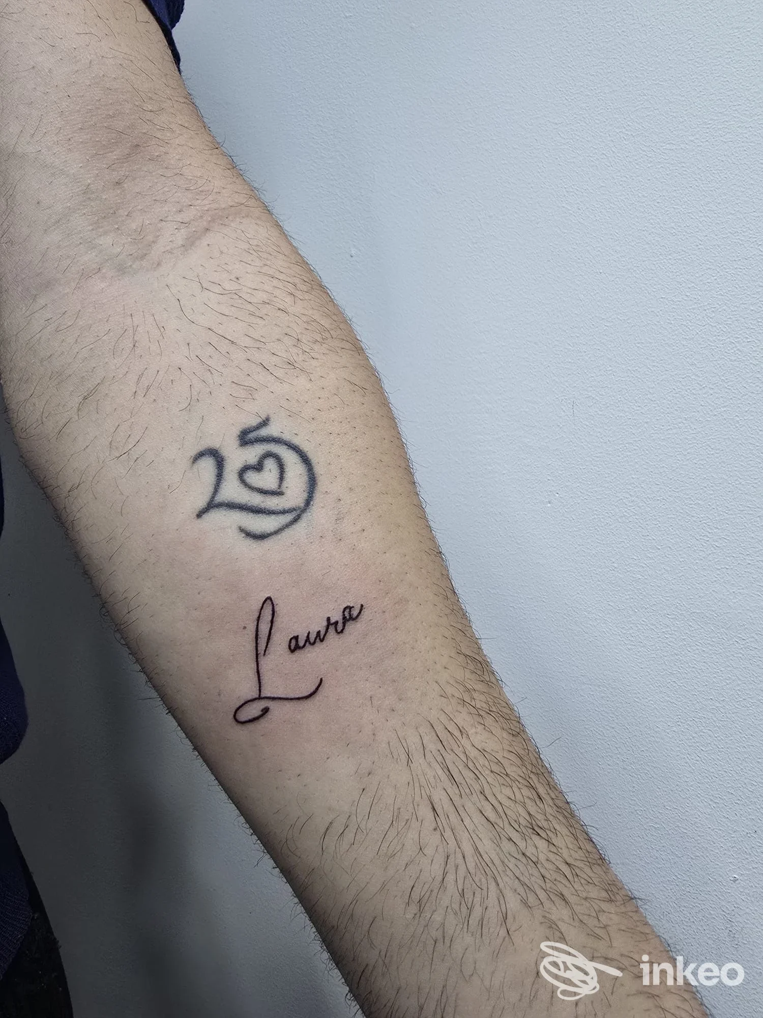 Avambraccio con tatuaggio visibile: testo stilizzato Lumo o Lulmo? e simbolo circolare scuro, pelle chiara, sfondo grigio. tatuaggio reale sul braccio.