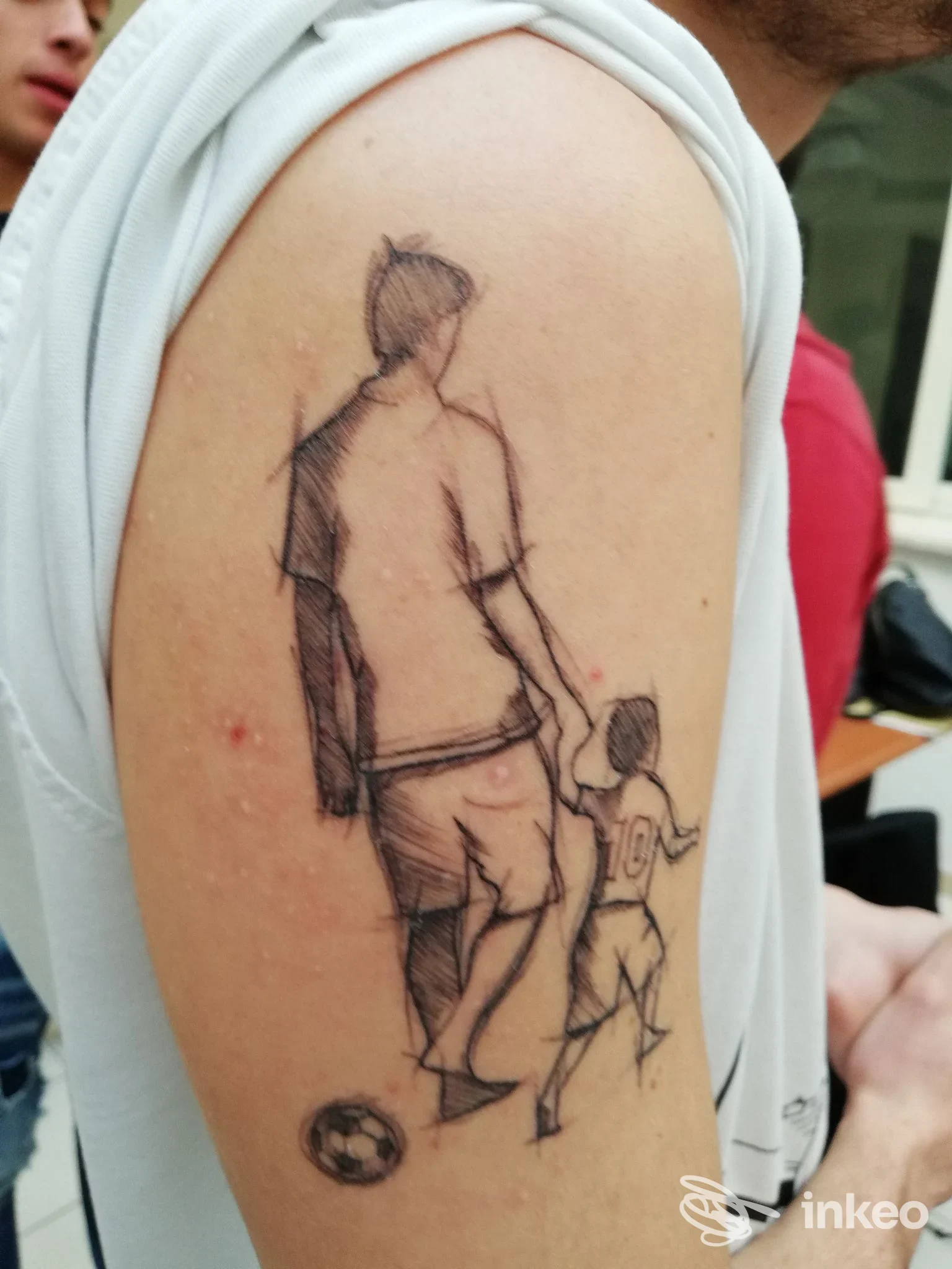 Tattoo sul braccio raffigurante due figure stilizzate, adulto e bambino, con pallone vicino; tattoo su pelle reale, scuro e con linee di schizzi, visibile su una parte del braccio.