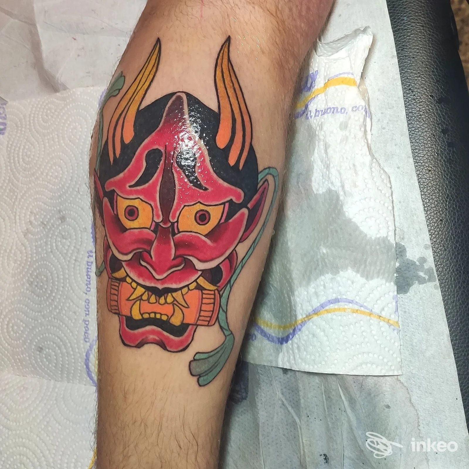 Tattoo colorato di una maschera demoniaca rossa con corna e dettagli neri e gialli sull'avambraccio, eseguito in uno studio; sfondo carta assorbente.