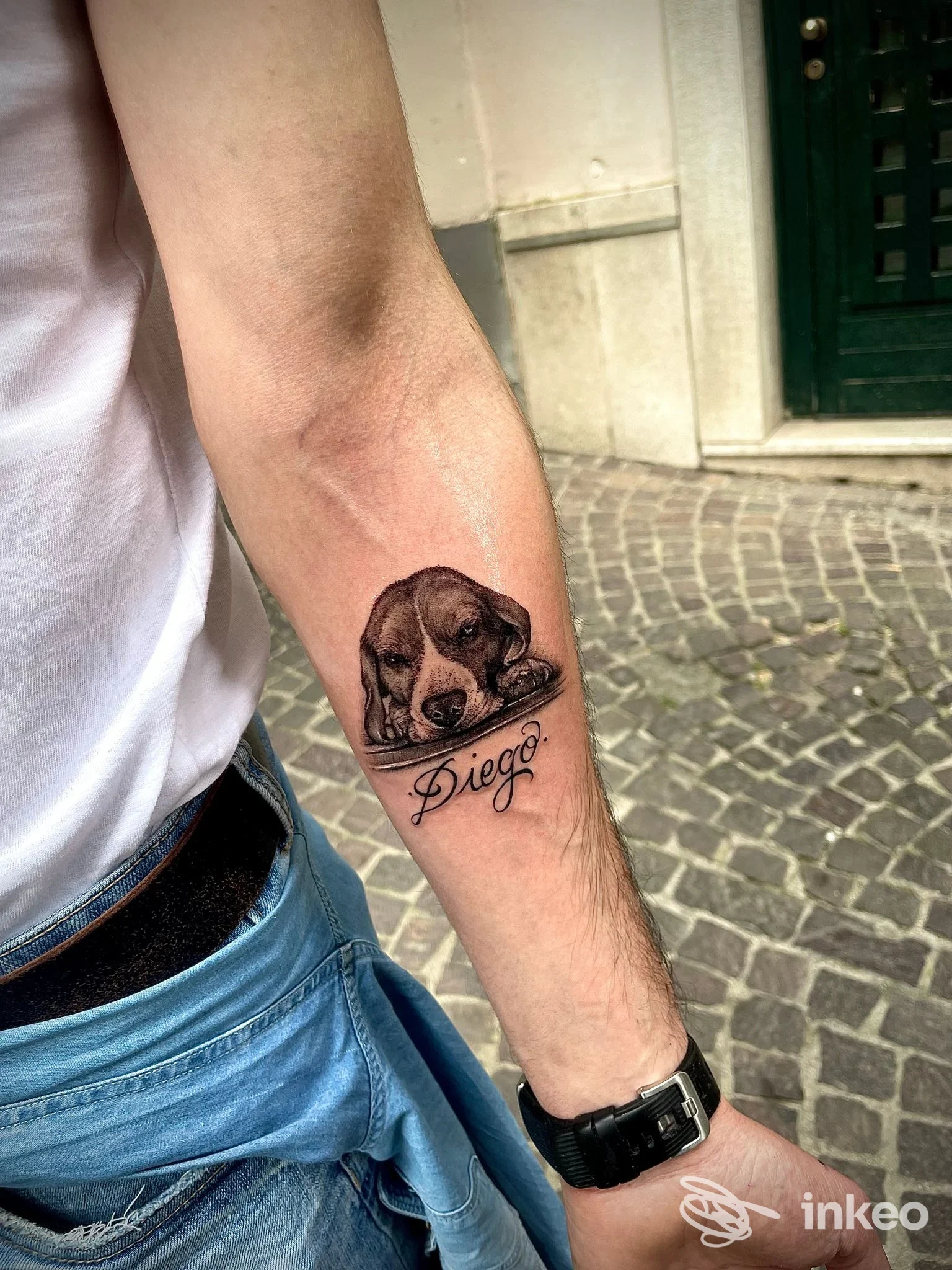 Tattoo visibile sul ventre dell’avambraccio sinistro: ritratto di cane scuro con nome Diego scritto sotto, eseguito su pelle reale; sfondo urbano in pietra e abbigliamento casual. Studio Ink Garage, Benevento