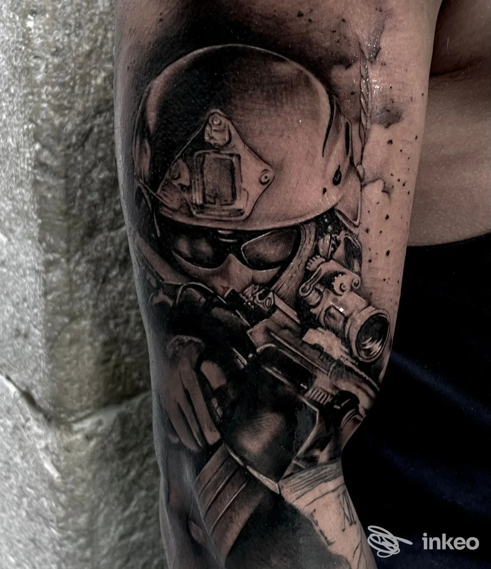 Tattoo realistico in bianco e nero su braccio: scena di un personaggio o equipaggiamento militare con dettaglio di elmetto, occhiali e arma.