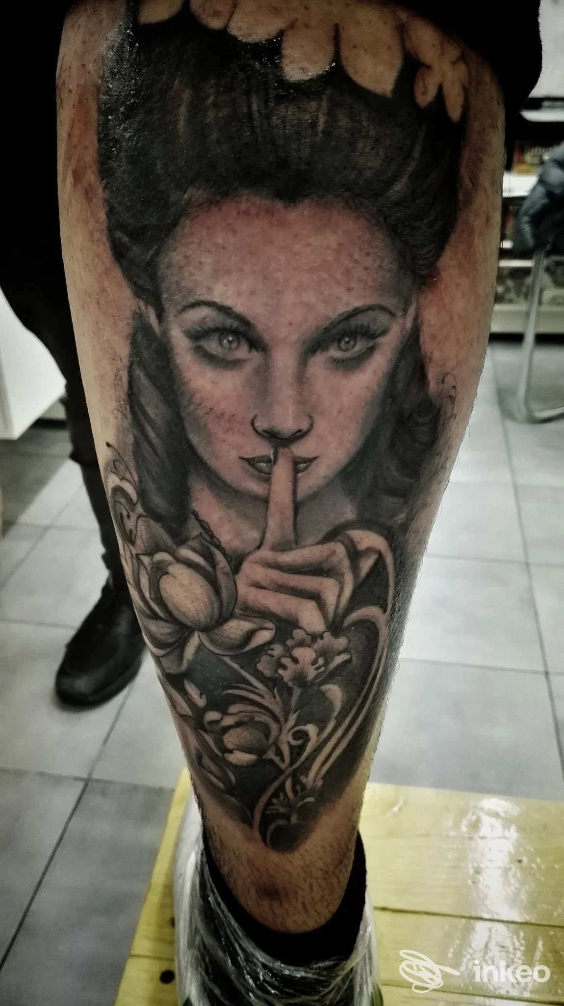 Tattoo raffigurante una donna con dito sull indice delle labbra, realizzata sulla gamba, con dettagli in bianco e nero e ornamenti floreali circostanti. Studio SUNINK TATTOO e Piercing, Pompei.