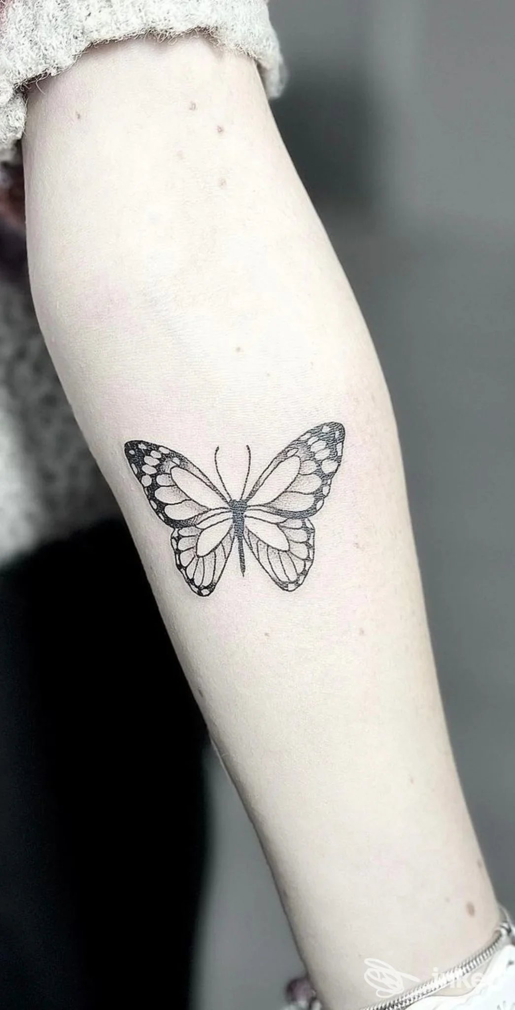 Tattoo a farfalla in bianco-neri su avambraccio, realizzato direttamente sulla pelle; sfondo neutro, nessun volto visibile.