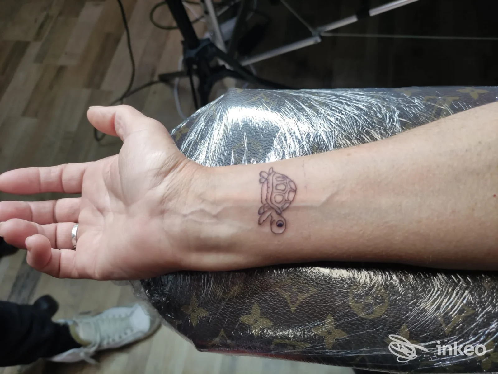 Tatuaggio reale sull’avambraccio sinistro: disegno in stile cartoon raffigurante una testa di scheletro, eseguito direttamente sulla pelle, visibile su sfondo tavolo da tattoo.