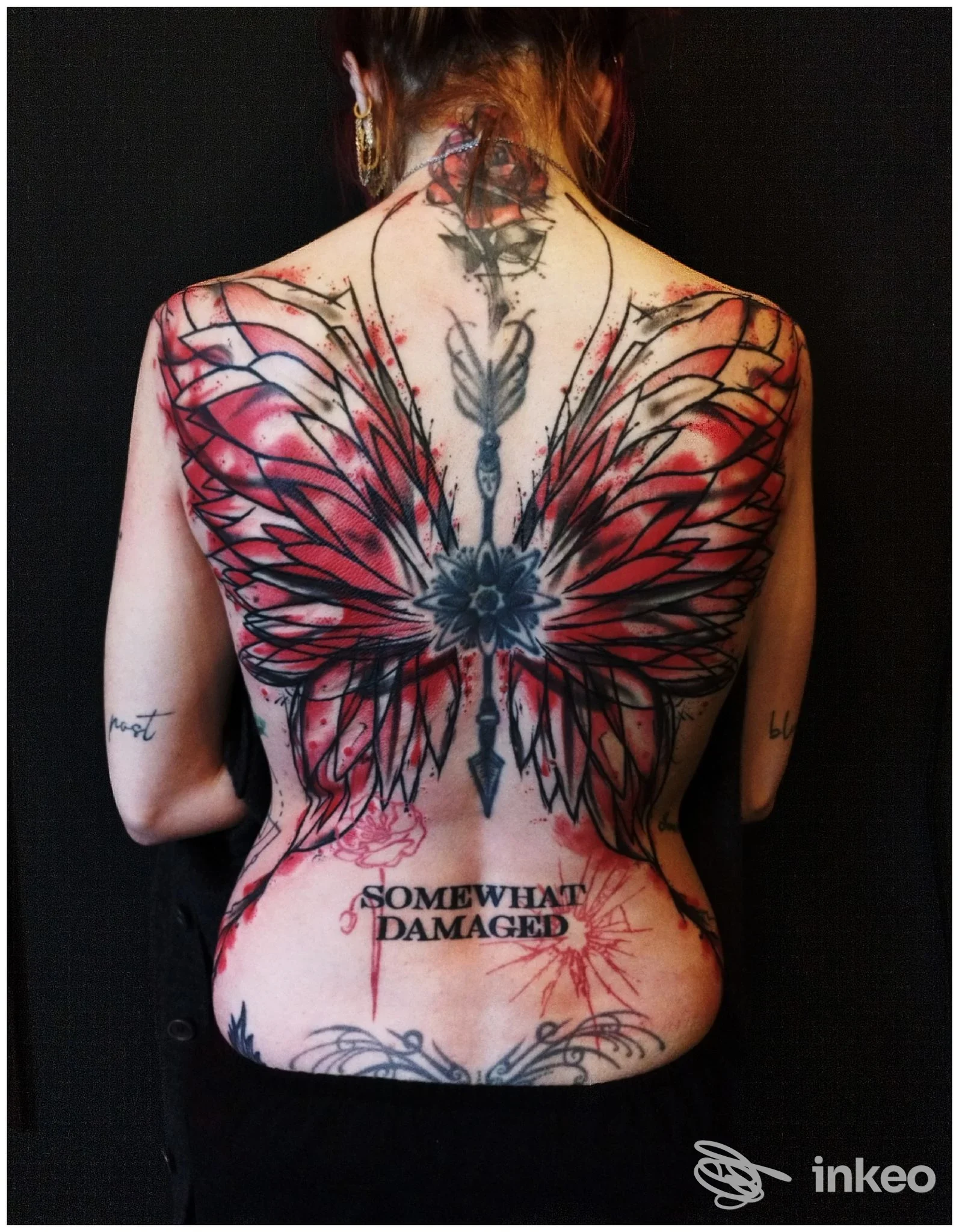 Tattoo reale sulla schiena: ali rosse e nere estese su tutta la schiena, con una linea centrale e un piccolo testo “SOMEWHAT DAMAGED” in basso, visibile su sfondo scuro. studio Black Tattoo, Caserta.