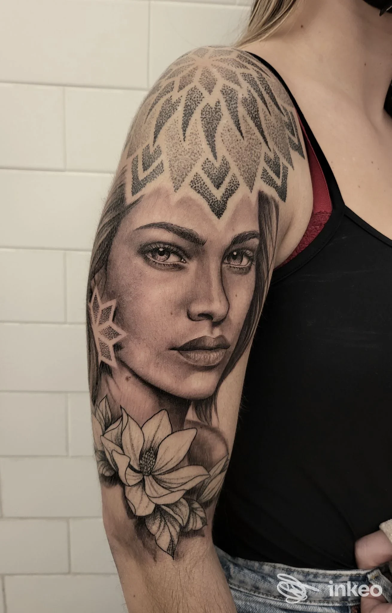 Braccio sinistro di una persona con tatuaggio realistico di volto femminile, ornamenti geometrici sul bicipite e un giglio sul braccio; sfondo con piastrelle bianche, attribuito allo studio Alessandro Sabatino Tattoo, Pontecagnano Faiano.