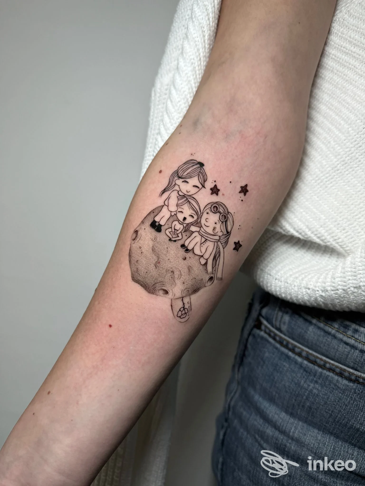 Tatuaggio su avambraccio raffigurante due personaggi su una luna, stile disegno cartone animato, circondato da stelle.