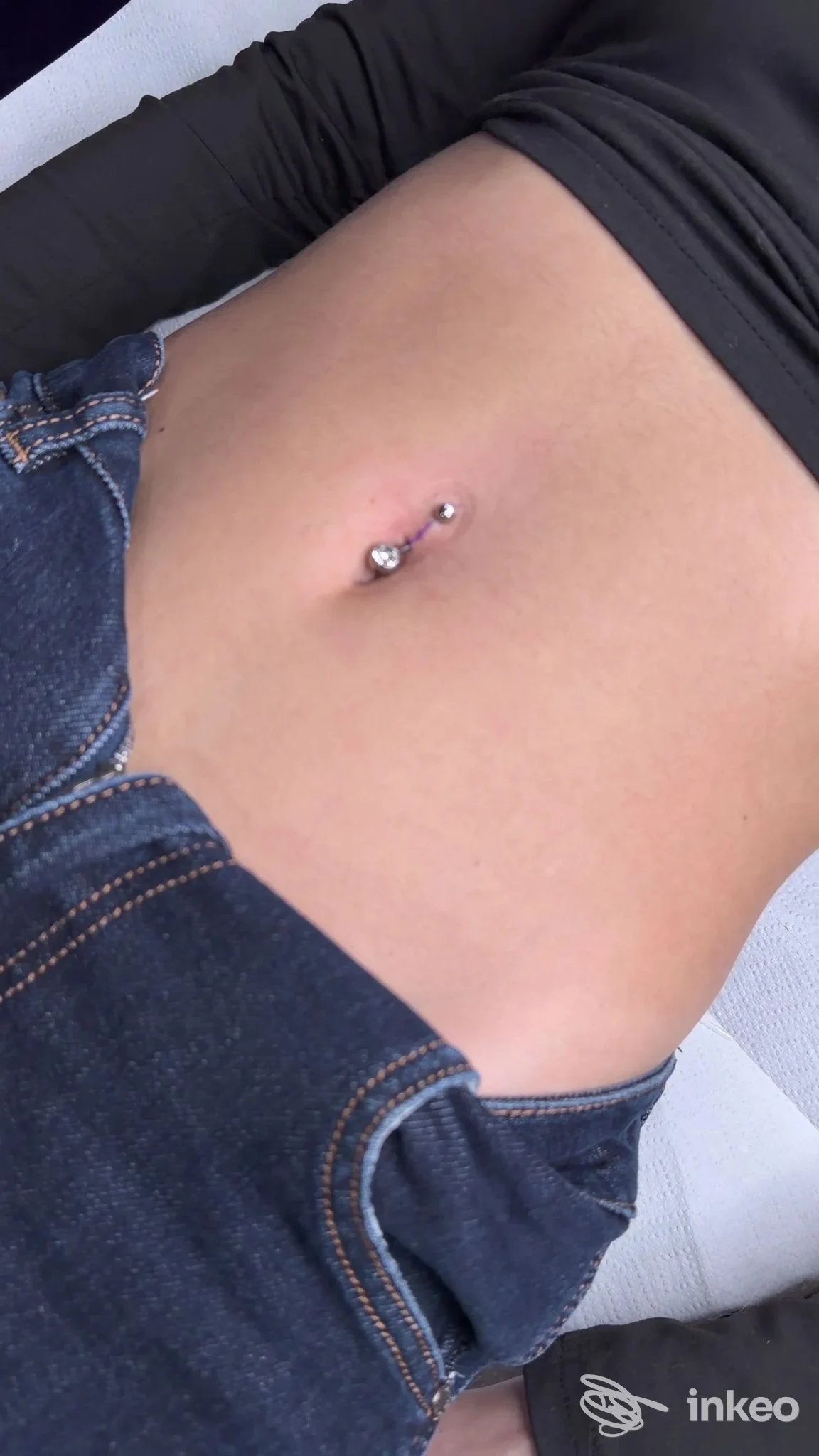 Piercing all'ombelico visibile sulla pelle, con jeans e top scuro, senza volto visibile.