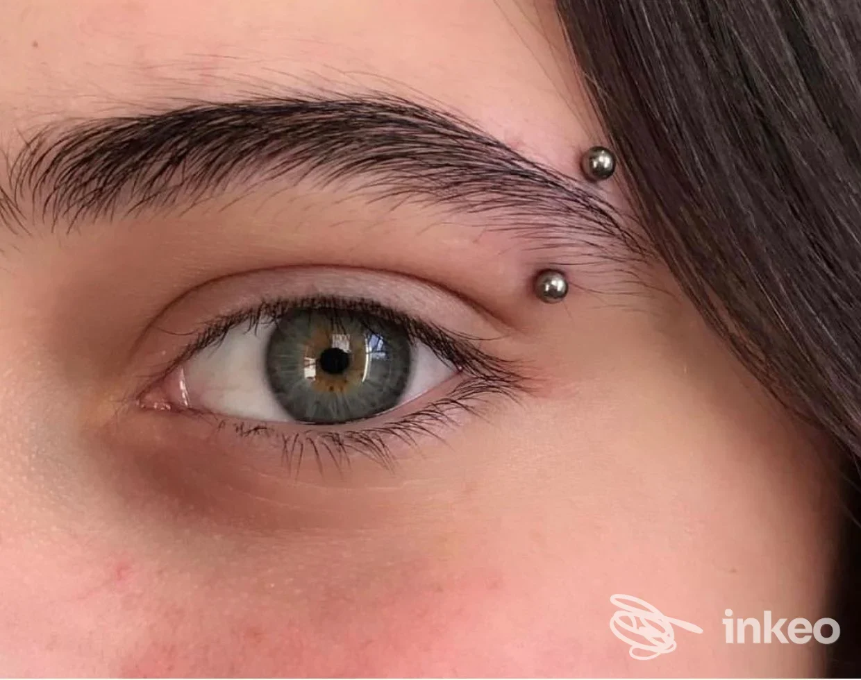 Primo piano sull’occhio sinistro: due piercing dermali posizionati sull’arcata sopraccigliare, visibili sulla pelle vicino all’occhio, senza mostrare l’intero volto.