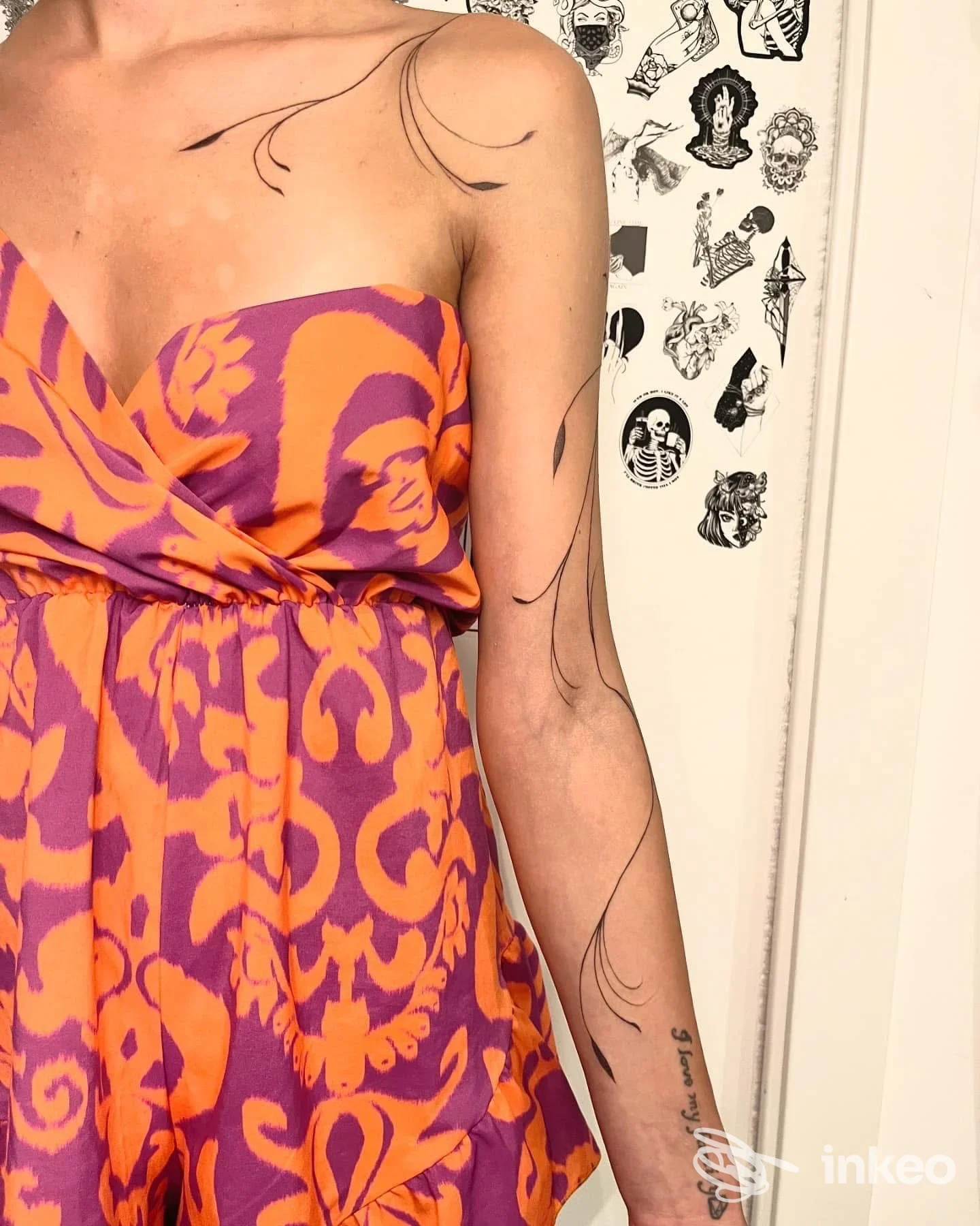 Una persona con abito viola e arancione mostra tatuaggi lineari e decorativi lungo la spalla e il braccio sinistro, visibili sulla pelle.