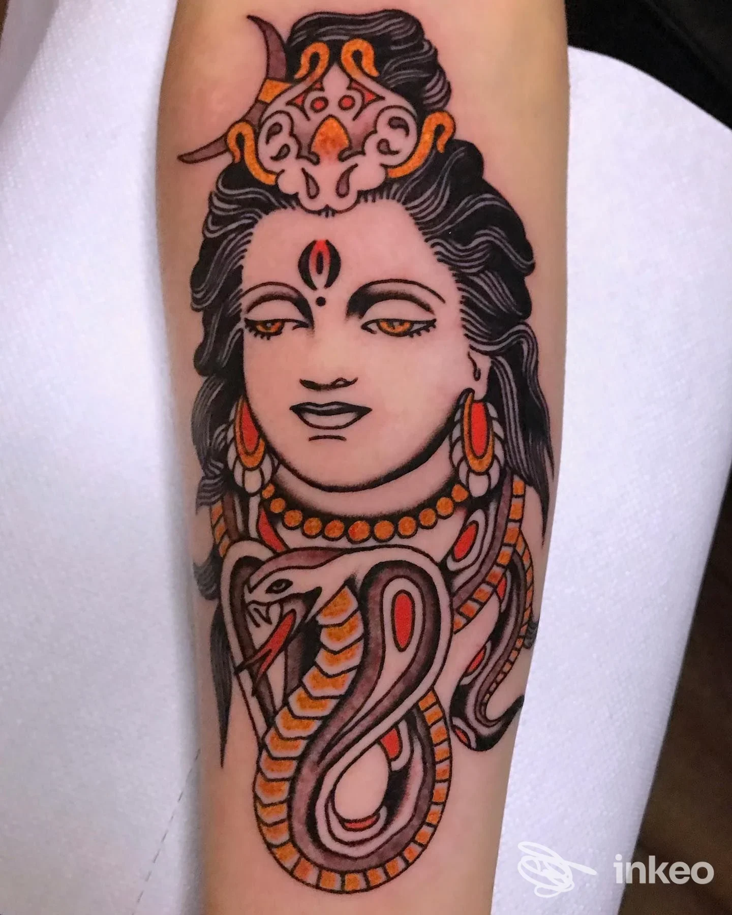 Tatuaggio su pelle raffigurante una divinità con serpenti: linee nere compensate con dettagli arancioni e rossi, viso sereno della divinità, ornamenti dorati; braccio come supporto del design; nessun overlay o testo esterno visibile.