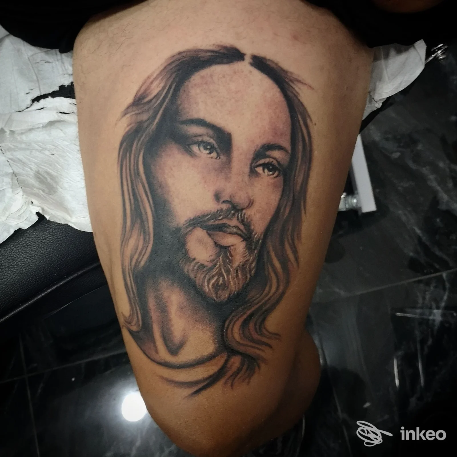 Tattoo sul fianco della gamba raffigurante un volto di Gesù, capelli lunghi e barba, eseguita con tratto realistico in bianco e nero su pelle chiara.