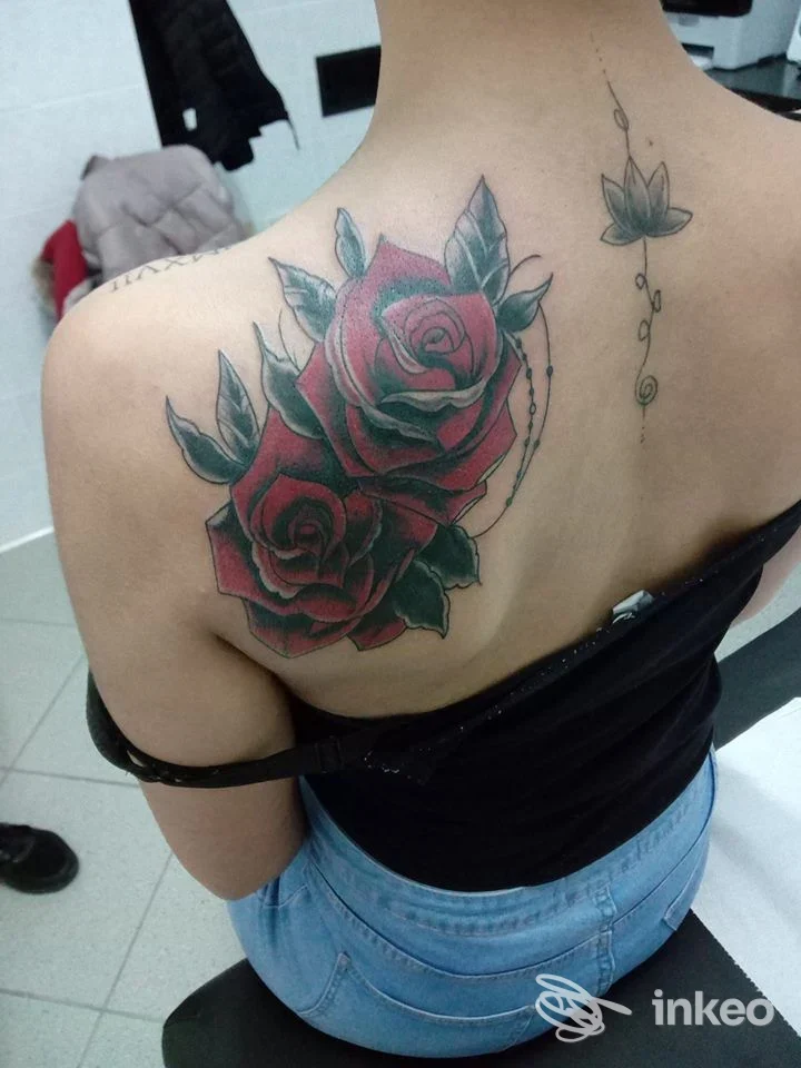Tattoo reale sulla schiena alta visibile, raffigurante rose rosse con foglie verdi e contorni neri, posizionato tra spalla e scapola sinistra; stile realistico/neo-tradizionale.