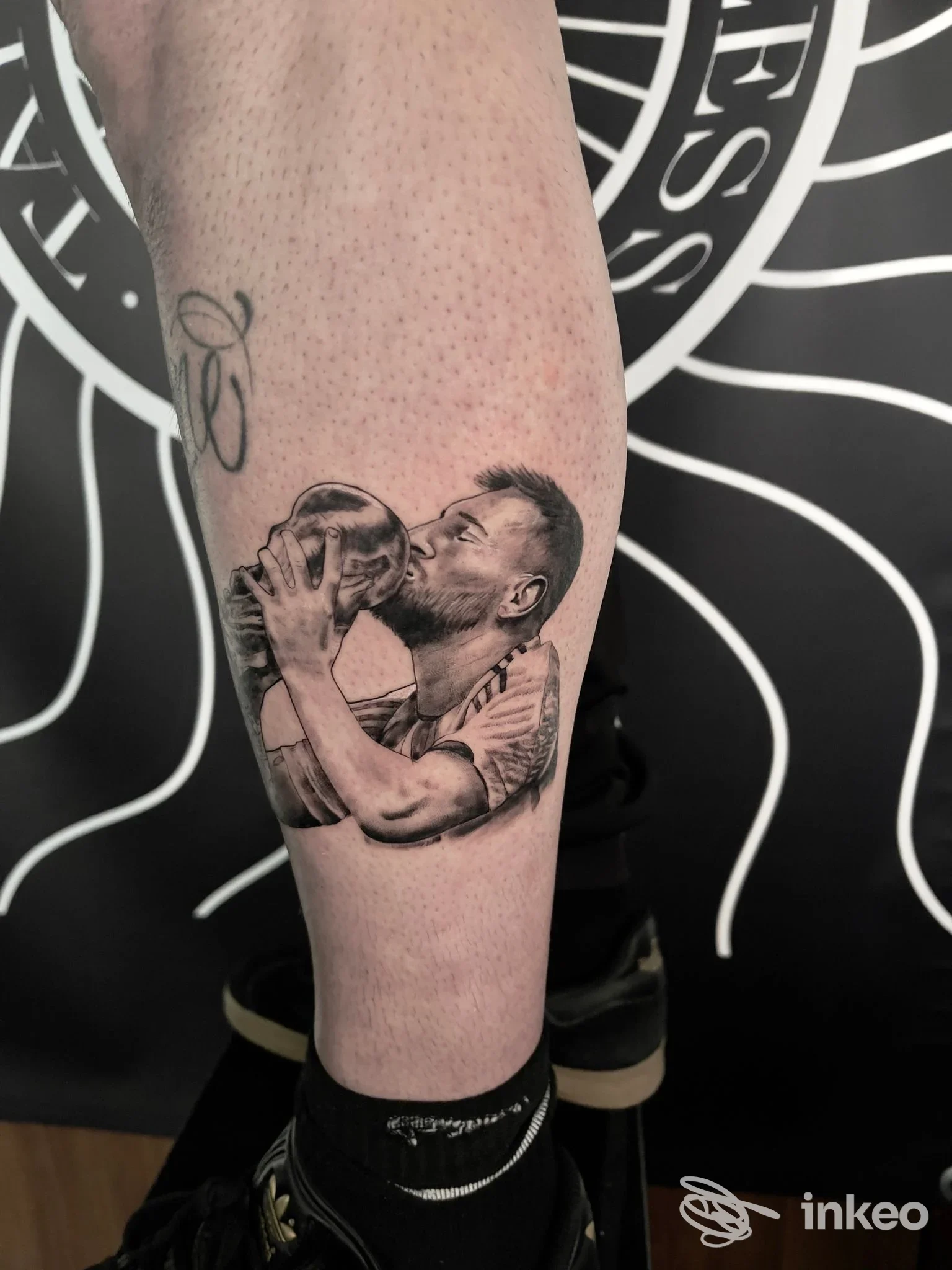Tattoo sul polpaccio raffigurante un uomo che tiene e sembra baciare una palla, realizzata sulla pelle senza elementi visivi di volto intero. sfondo nero con grafica bianca.