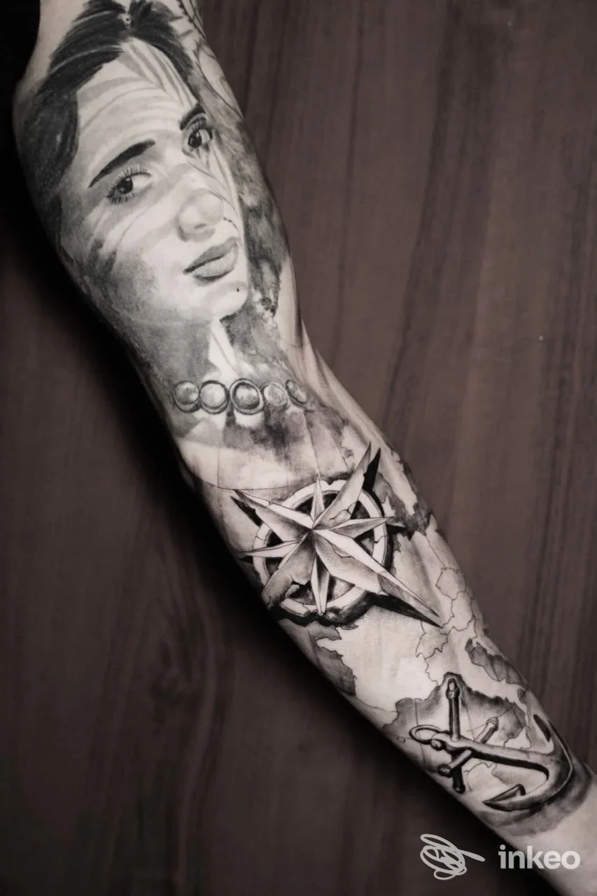 Braccia tatuata con ritratto realistico di una donna, accompagnata da una bussola e ancora in stile bianco e nero; braccio coperto da tattoo dal polso al gomito.