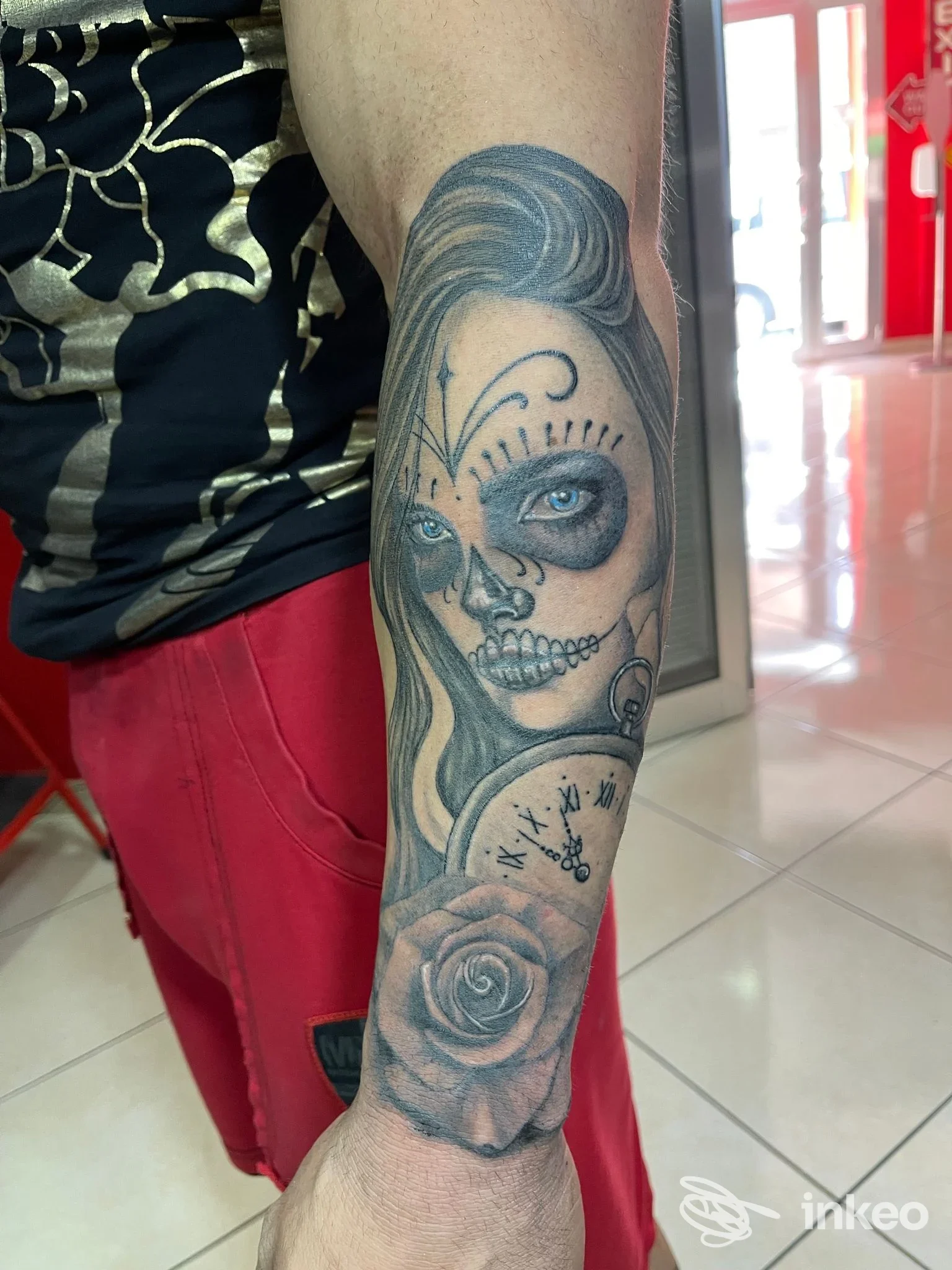 Tattoo visibile sul braccio: ritratto stilizzato dalla testa di una donna con elementi Day of the Dead, rose e dettagli ornamentali, eseguito su pelle chiara.