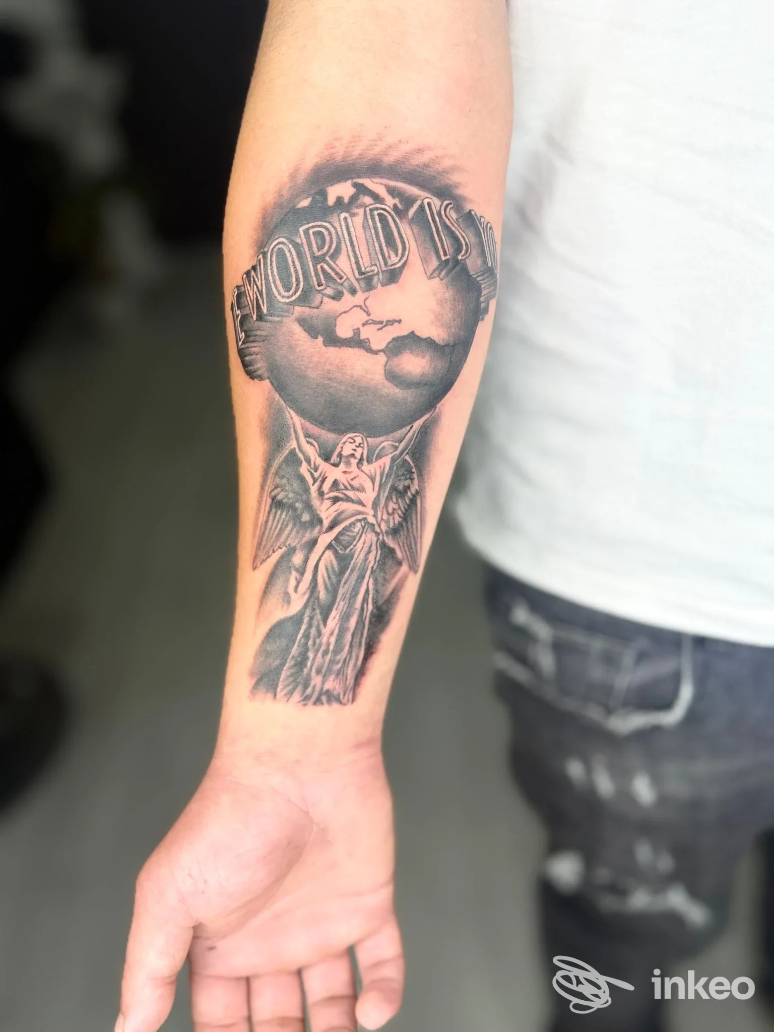 Avambraccio di una persona con tatuaggio realistico: una scena con globo terrestre e figura angelica in tonalità nero/grigio, testo World parzialmente visibile.