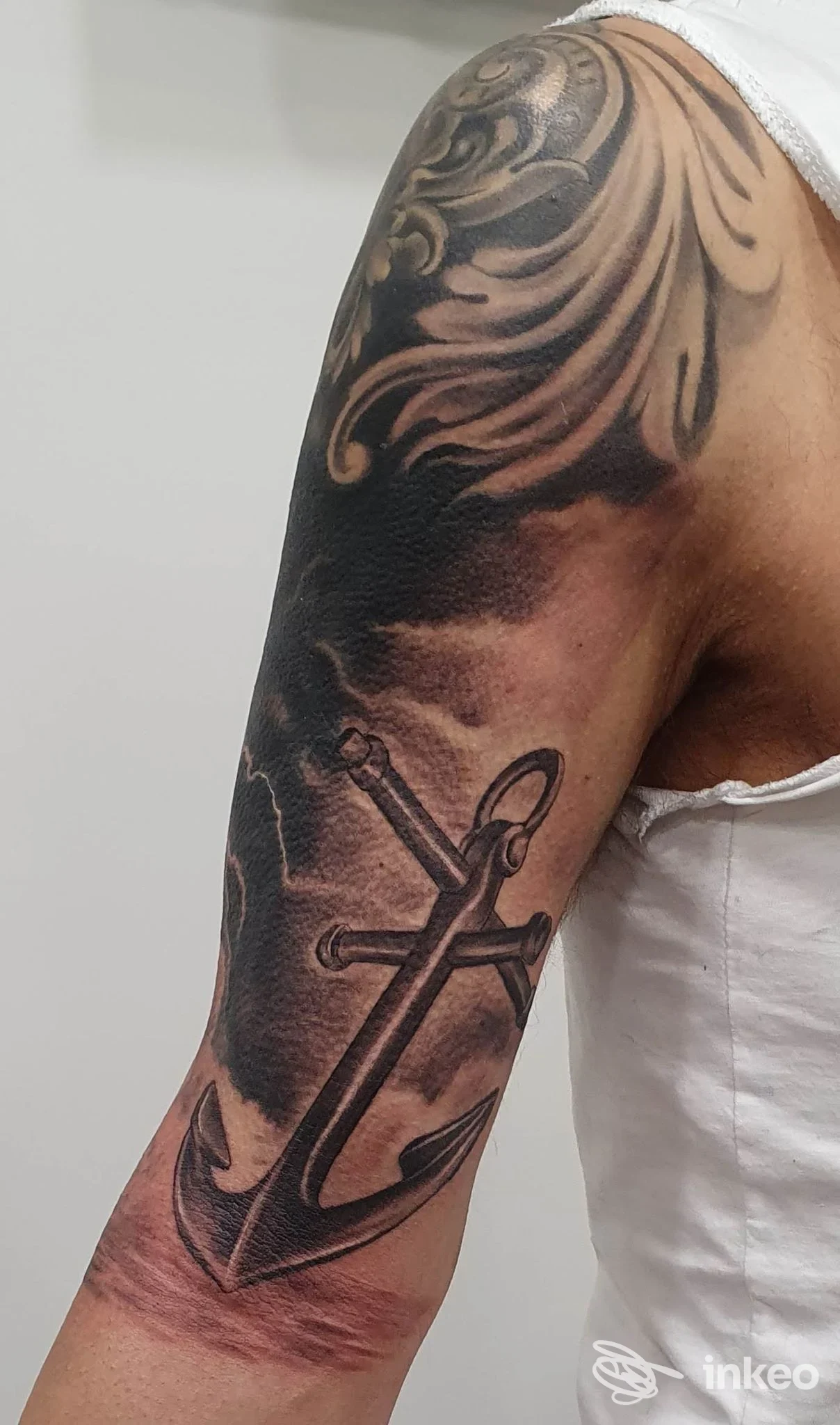 Un braccio visibile mostra un tatuaggio reale di ancora marina: pelle tratteggiata e sfumata, con dettagli dell'inchiostro ben definiti. Si vede solo una porzione del braccio, senza volto; lo sfondo è neutro. Studio Agostino Ferraiuolo Tattoo Store, Casavatore.