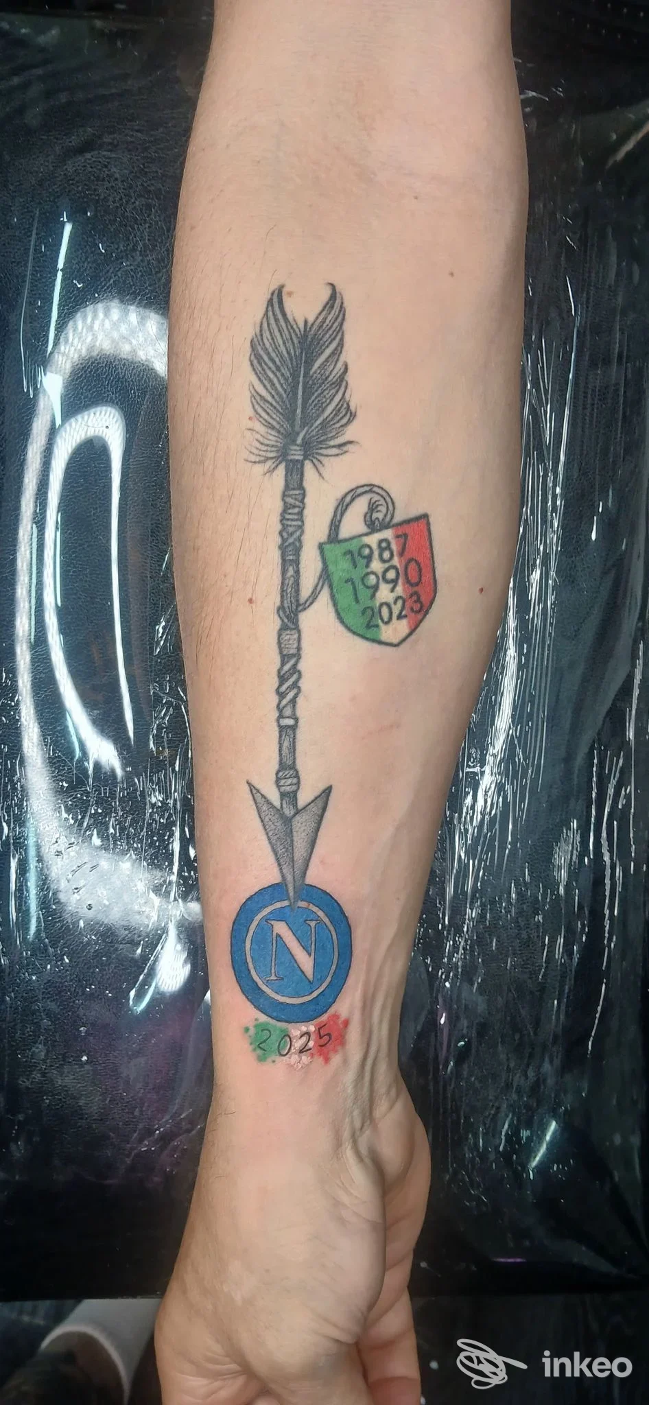 Tatuaggio visibile sul polso: freccia stylizzata con piuma e scudo colorato, su pelle chiara; colore principale nero/blu con dettagli verdi e rossi, realizzato su braccio.