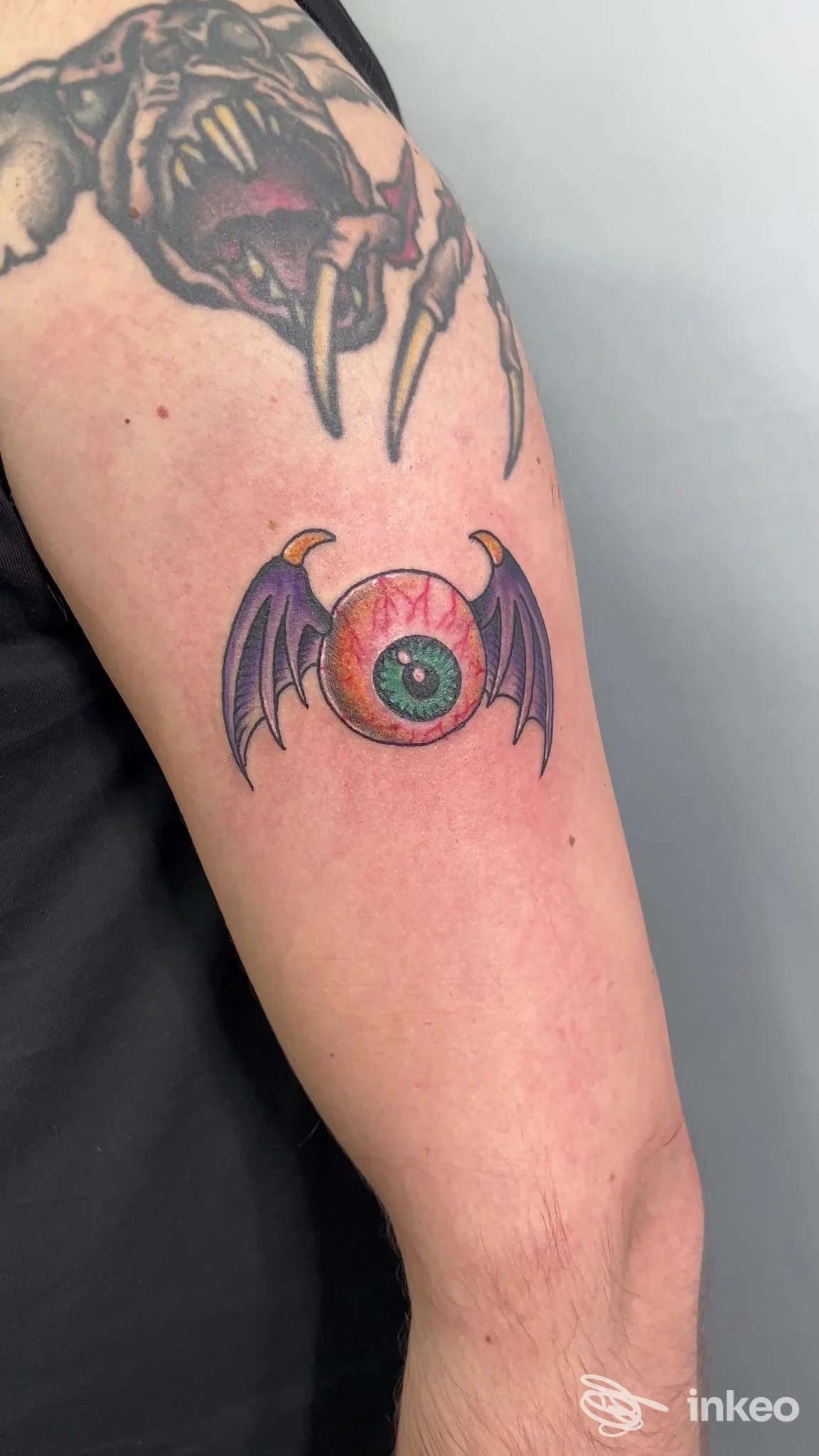Tatuaggio sul braccio: un globo rosso con un occhio verde al centro e ali da pipistrello viola, colorato e definito.