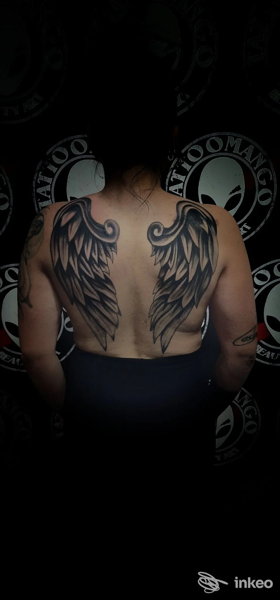 Tatuaggio reale di ali con dettagli scuri sulle scapole di una persona vista di schiena, sfondo con logo studio Tattoomango Napoli.