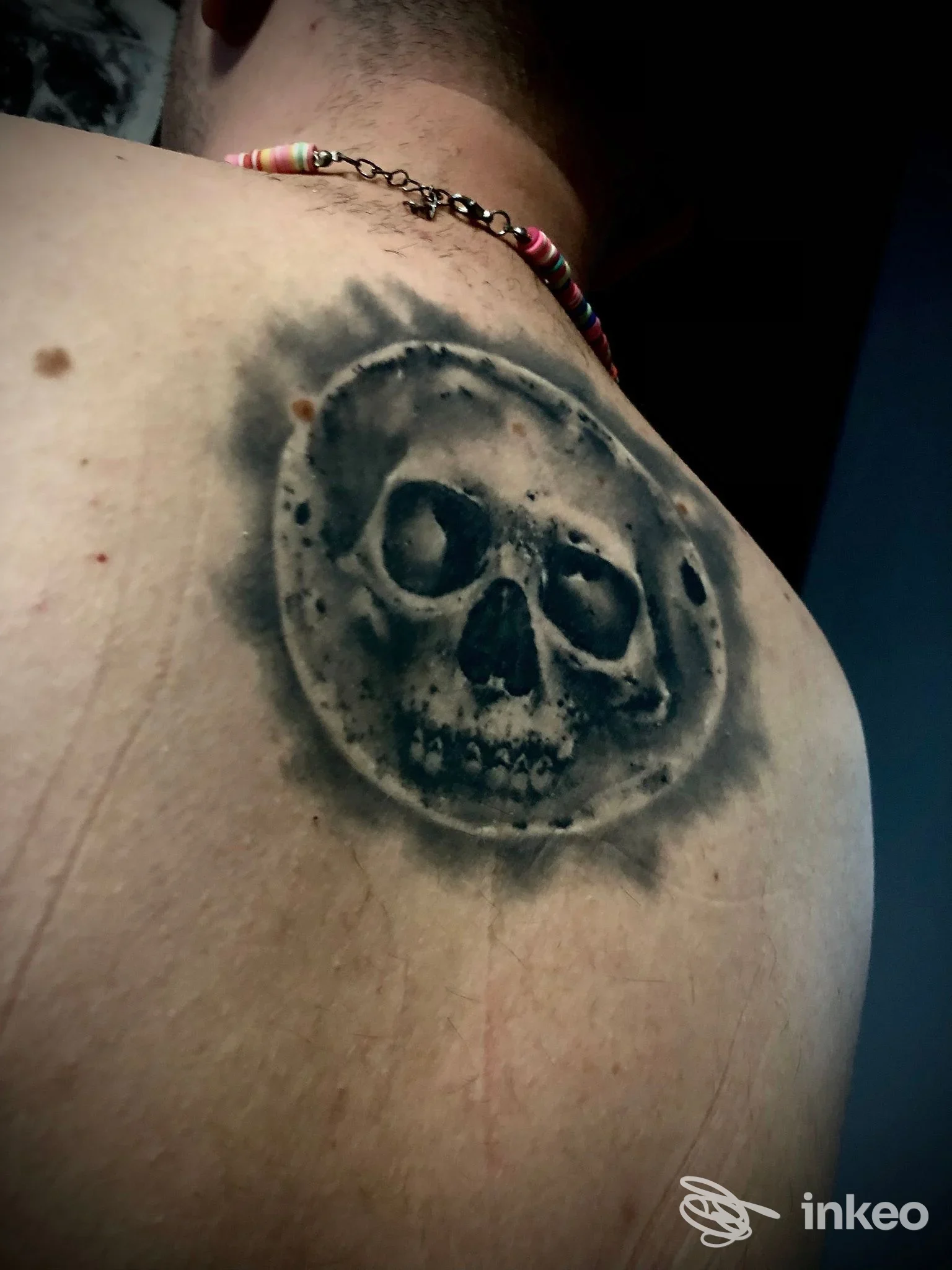Tattoo su spalla raffigurante un teschio circondato da un alone scuro, visibile su pelle chiara; collana colorata sullo sfondo.