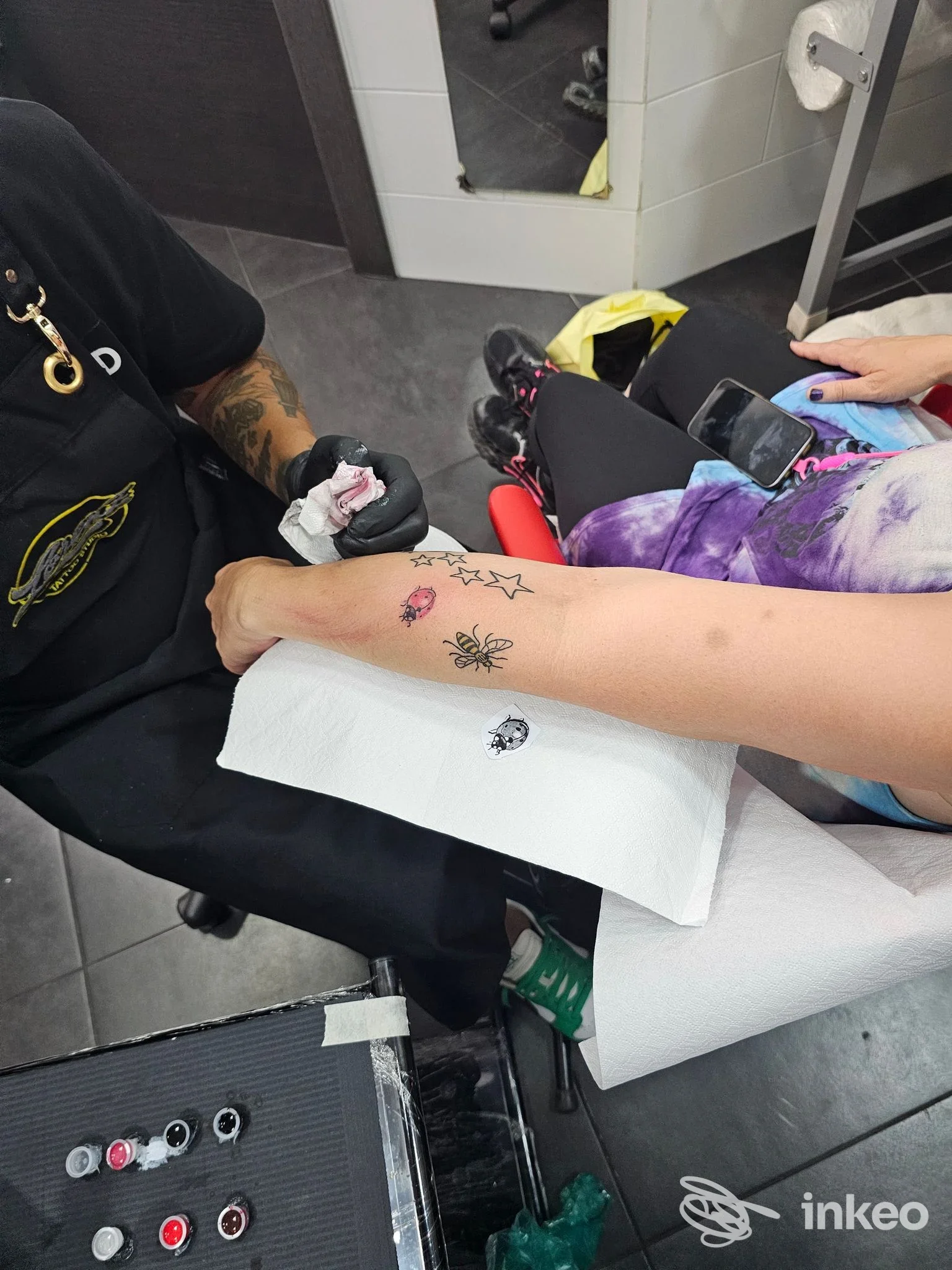 Primo piano su una gamba tatuata in studio, mentre il tatuatore lavora e avvolge la ferma con una salvietta; sullo sfondo si vedono strumenti e sedie da tatuaggio.