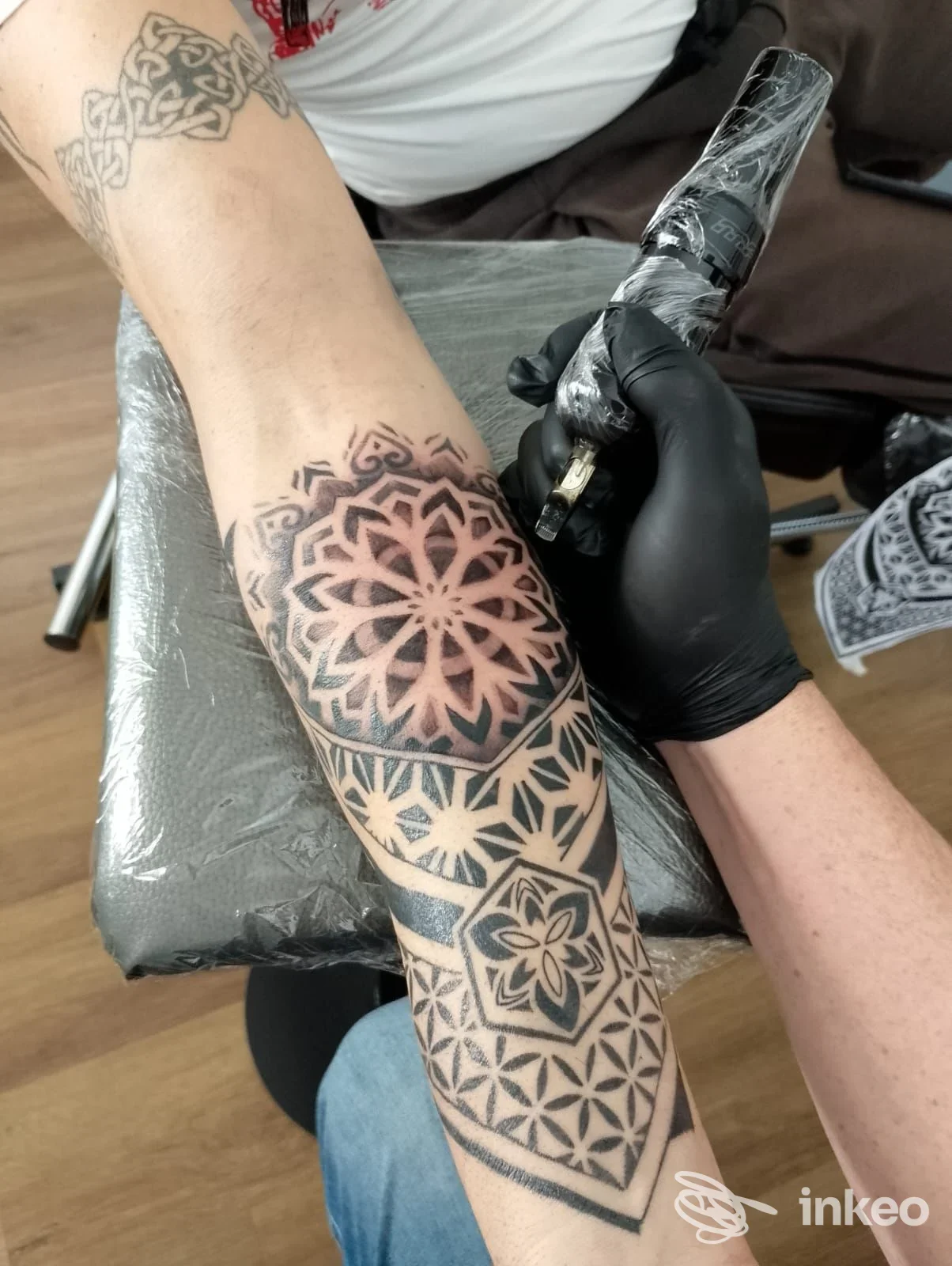 Un braccio con un grande tatuaggio geometrico in corso di realizzazione: l'artista indossa guanti neri e lavora con una macchina per tatuare sull'avambraccio.